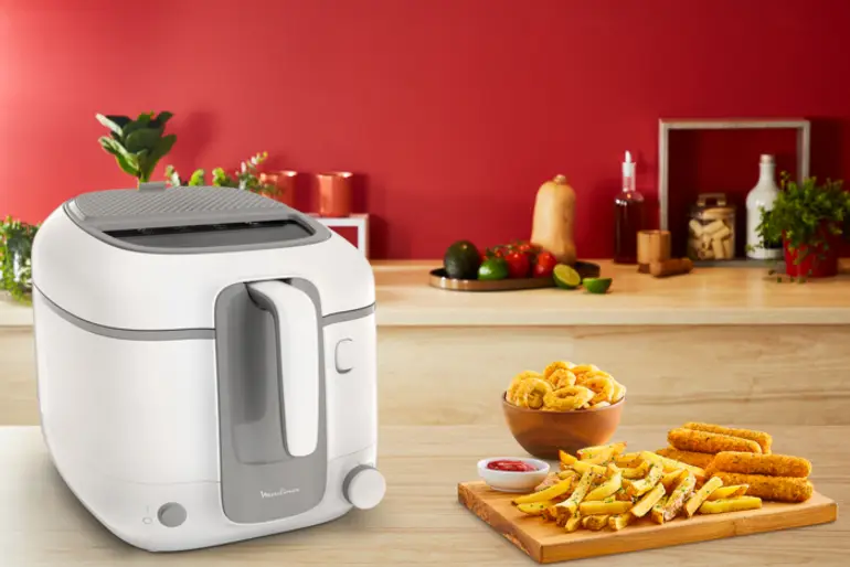 Witte en grijze friteuse, met eten en ingrediënten op een houten tafel en rode achtergrond.