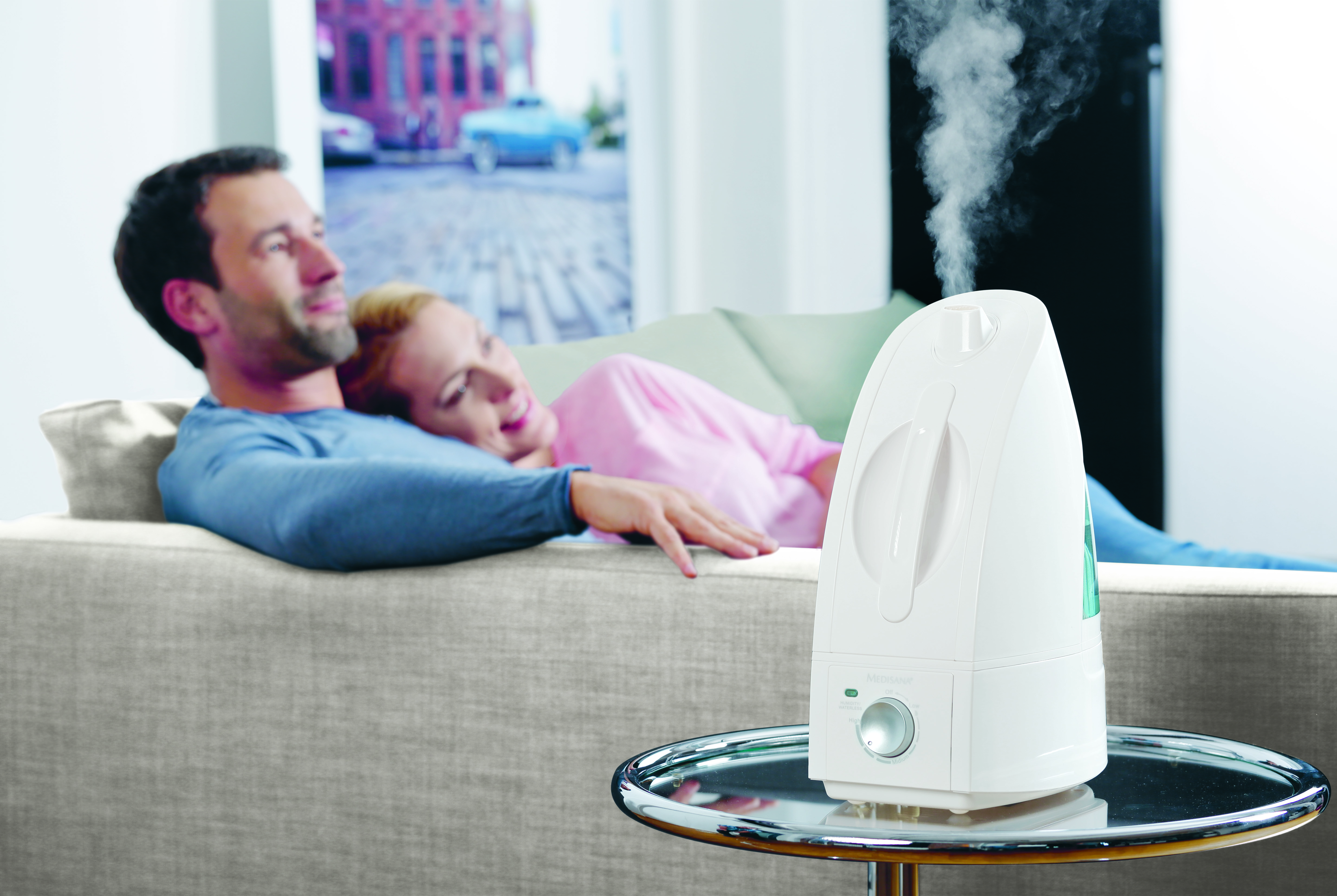 Un humidificateur sur une table avec de la vapeur, et un couple se relaxant sur un canapé en arrière-plan flou.