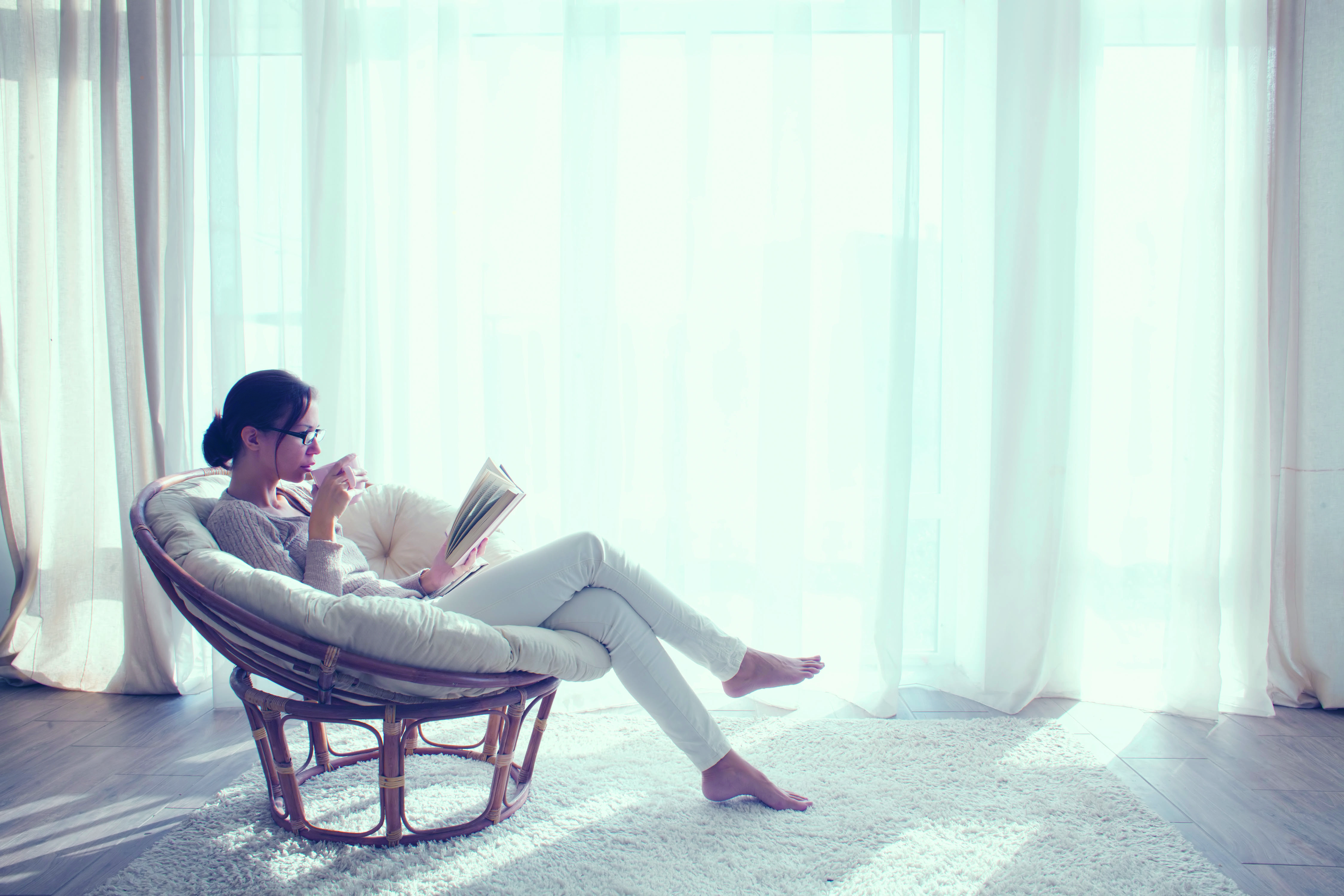 Femme lisant un livre dans un fauteuil près d'une fenêtre avec rideaux blancs. Soleil brille.
