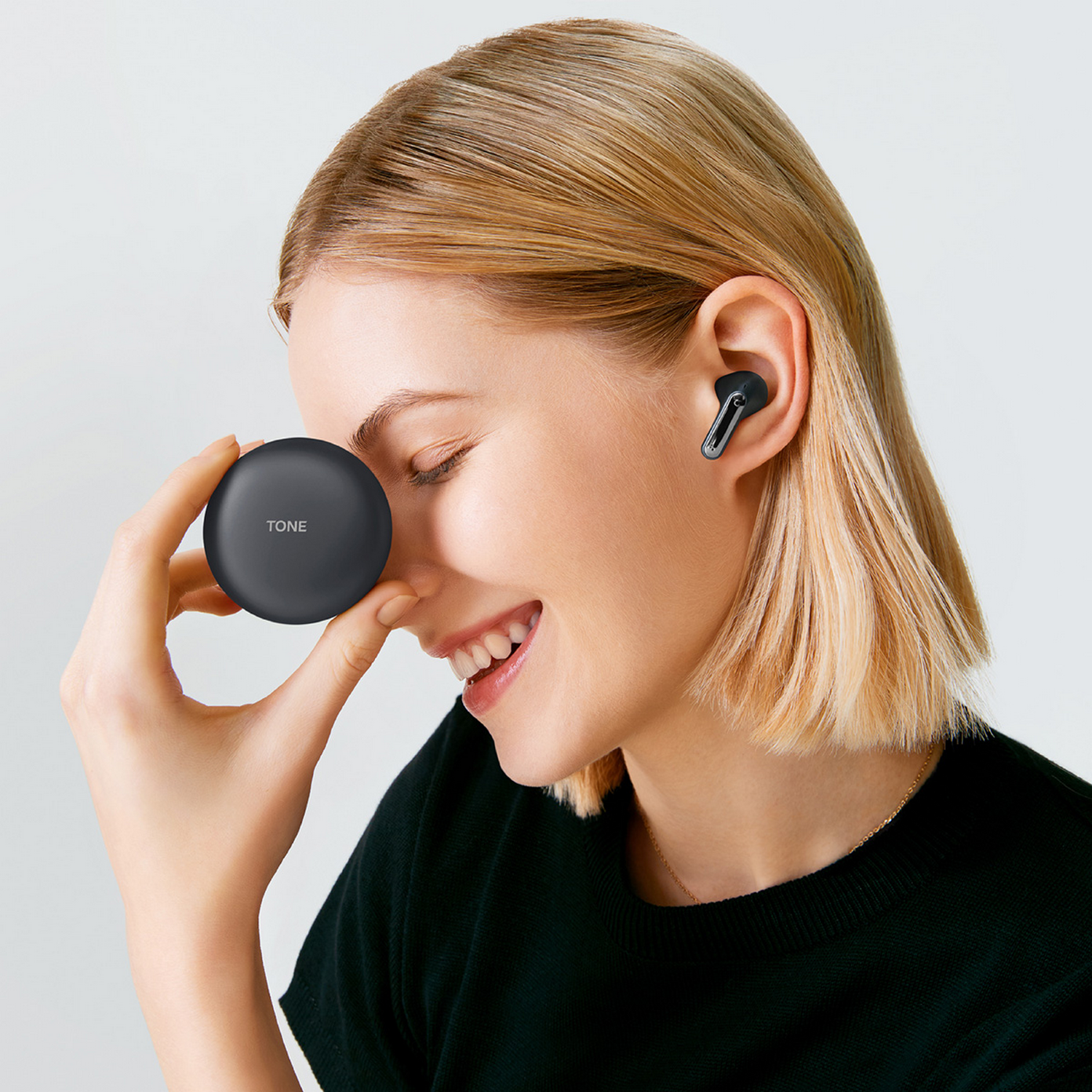 Mujer rubia sonriendo, sosteniendo un estuche negro de auriculares, un auricular en la oreja.