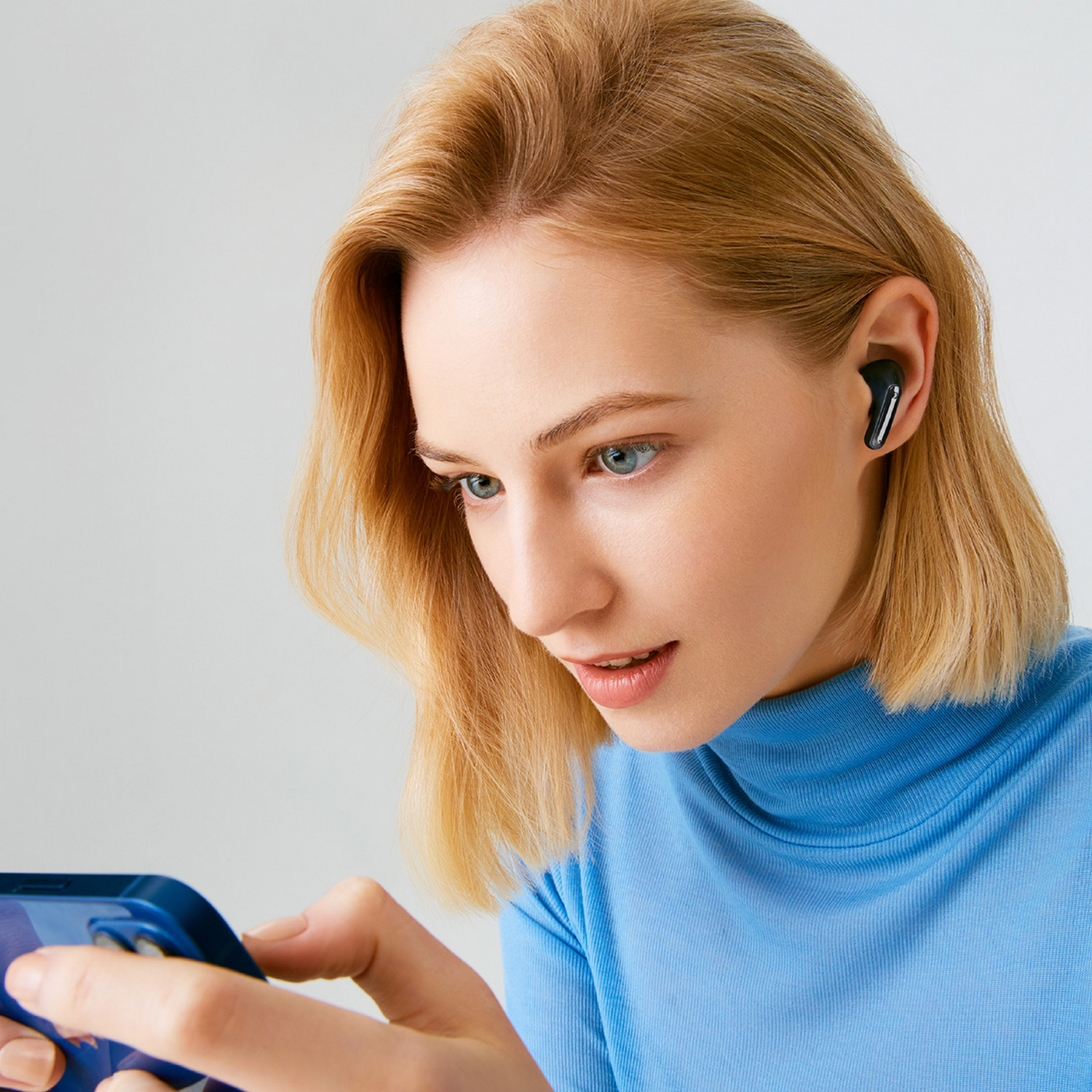 Una mujer con pelo rubio con una camisa azul usando un teléfono. Tiene auriculares negros en la oreja.