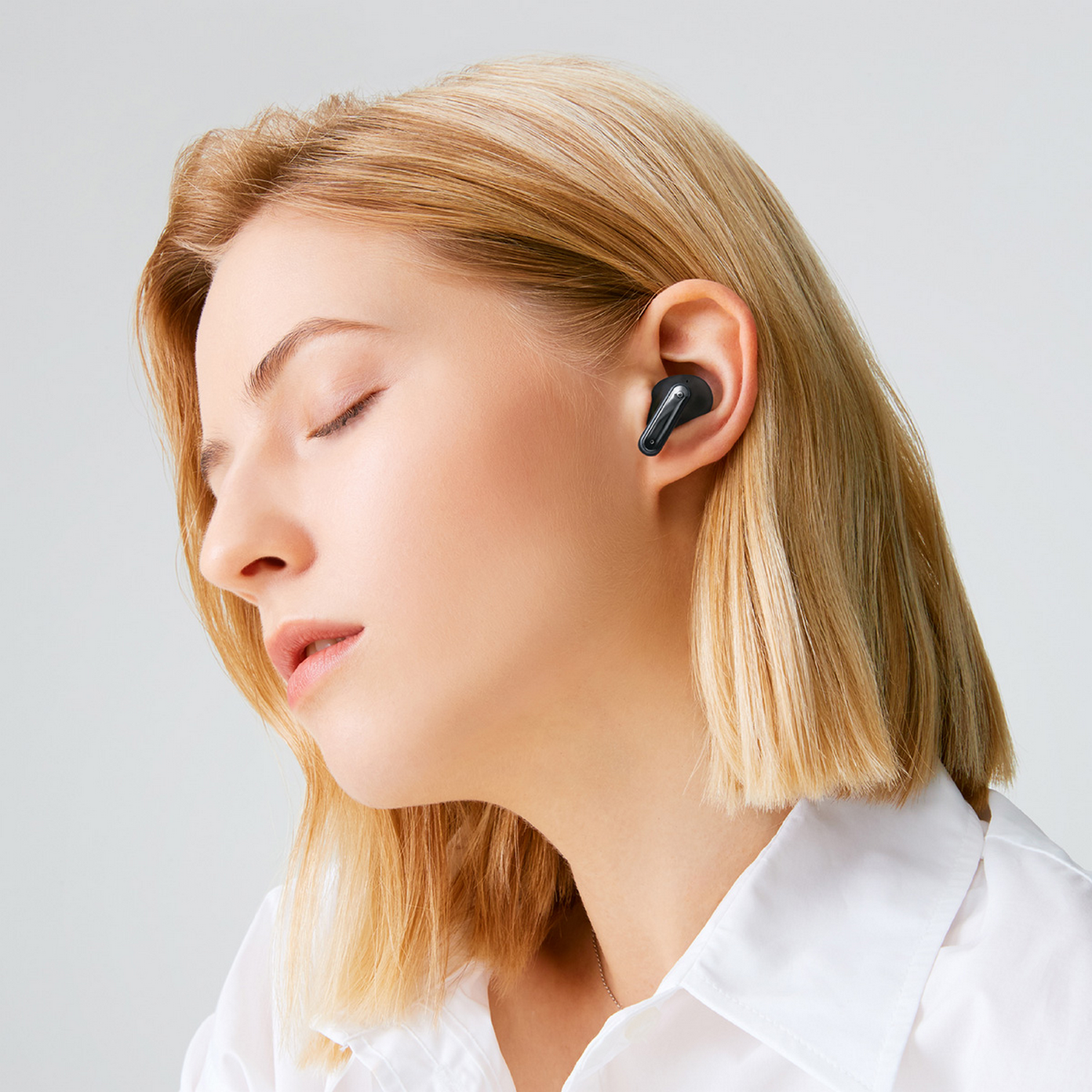 Mujer con cabello rubio usando un auricular negro en su oreja derecha, mirando hacia la izquierda sobre un fondo gris.