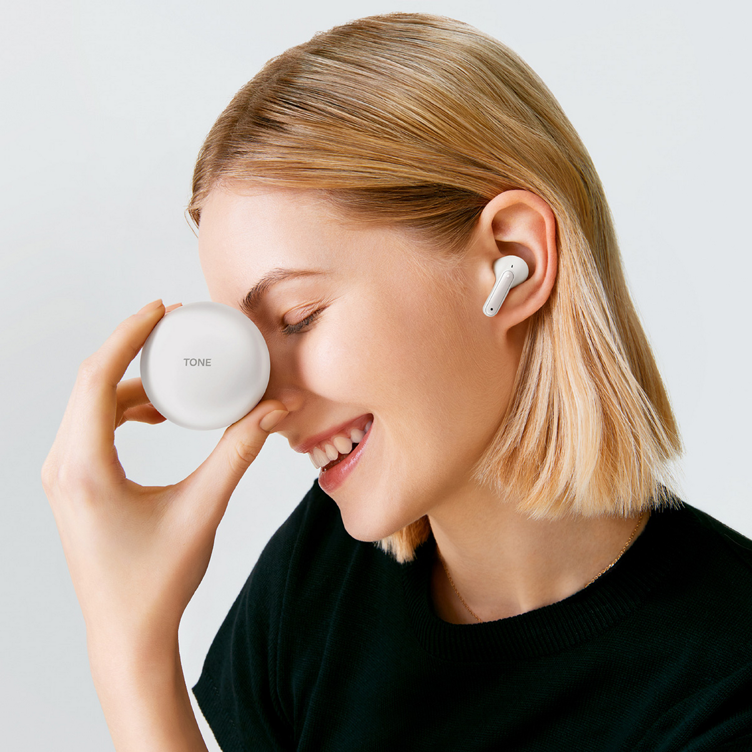 Mujer sonríe, sosteniendo un estuche blanco para auriculares con la etiqueta 'TONE' cerca de su ojo cerrado. Un auricular está en su oído. Fondo blanco.