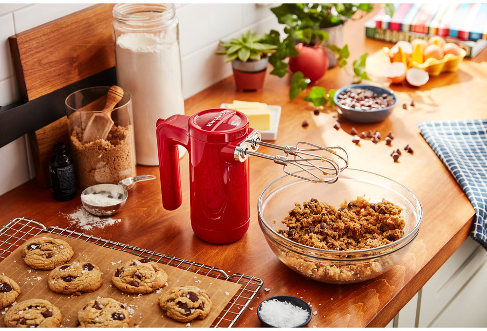 Red hand mixer, bowl with batter, cookies, and ingredients on a table.
