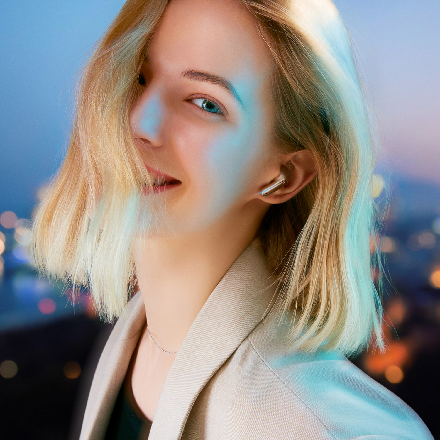 Mujer rubia sonriendo, con una chaqueta ligera y auriculares. Luces de la ciudad al fondo.