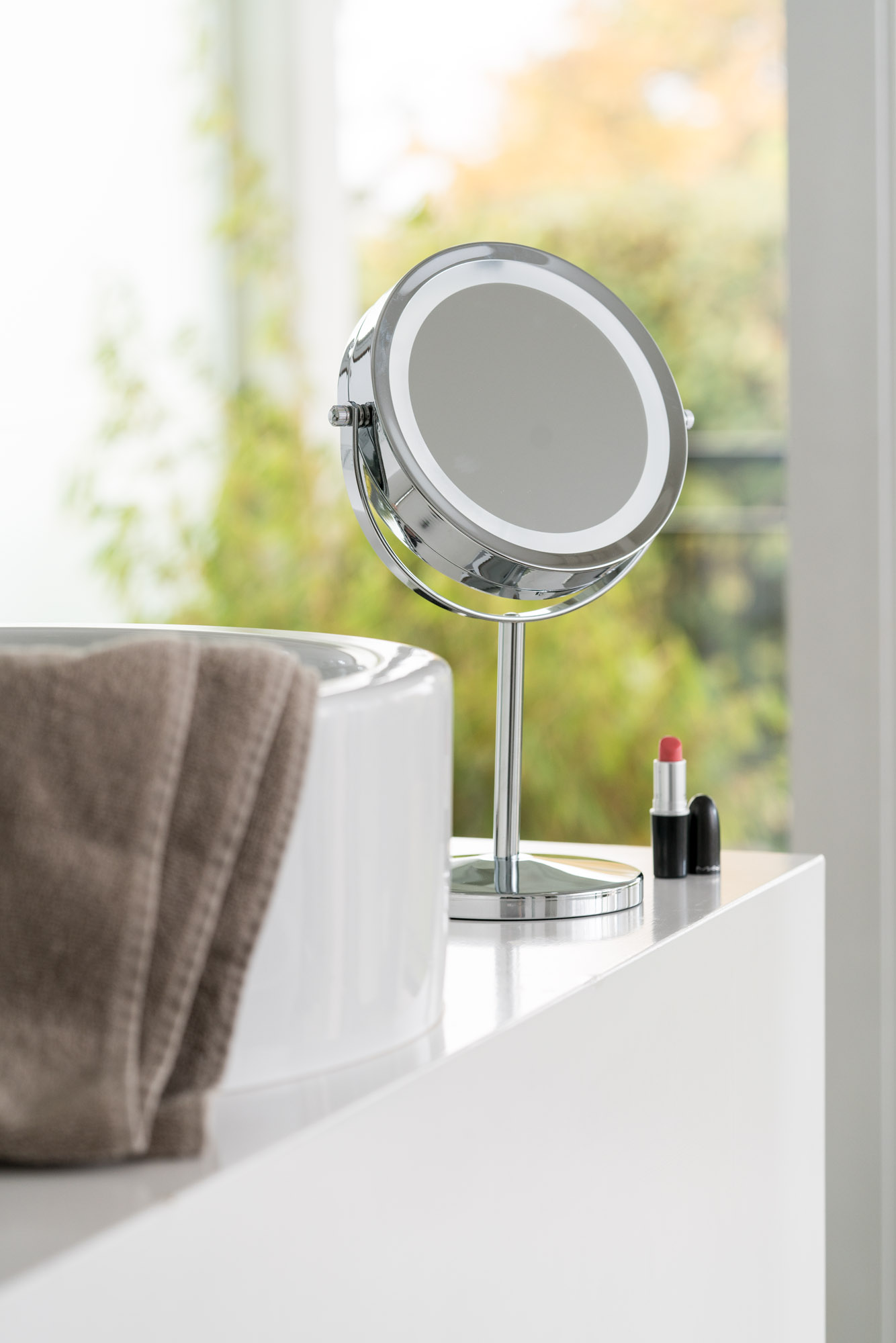 A chrome mirror, lipstick, and a towel rest on a white surface. The background is a window with greenery.