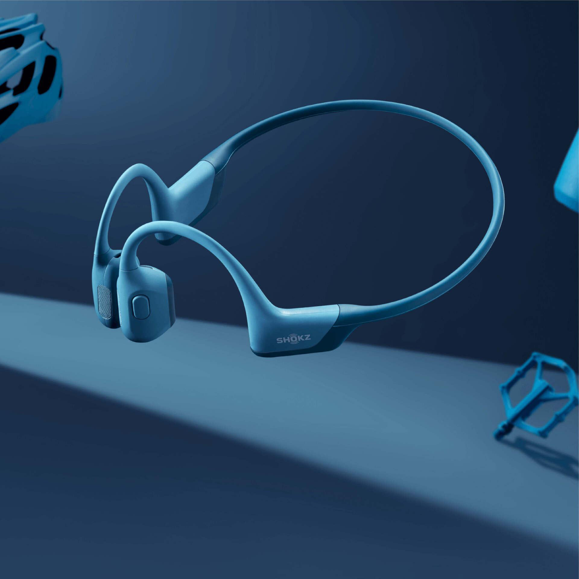 Blaue Shokz-Kopfhörer, Fahrradhelm und Pedal auf blauem Hintergrund. Die Kopfhörer sind im Fokus.