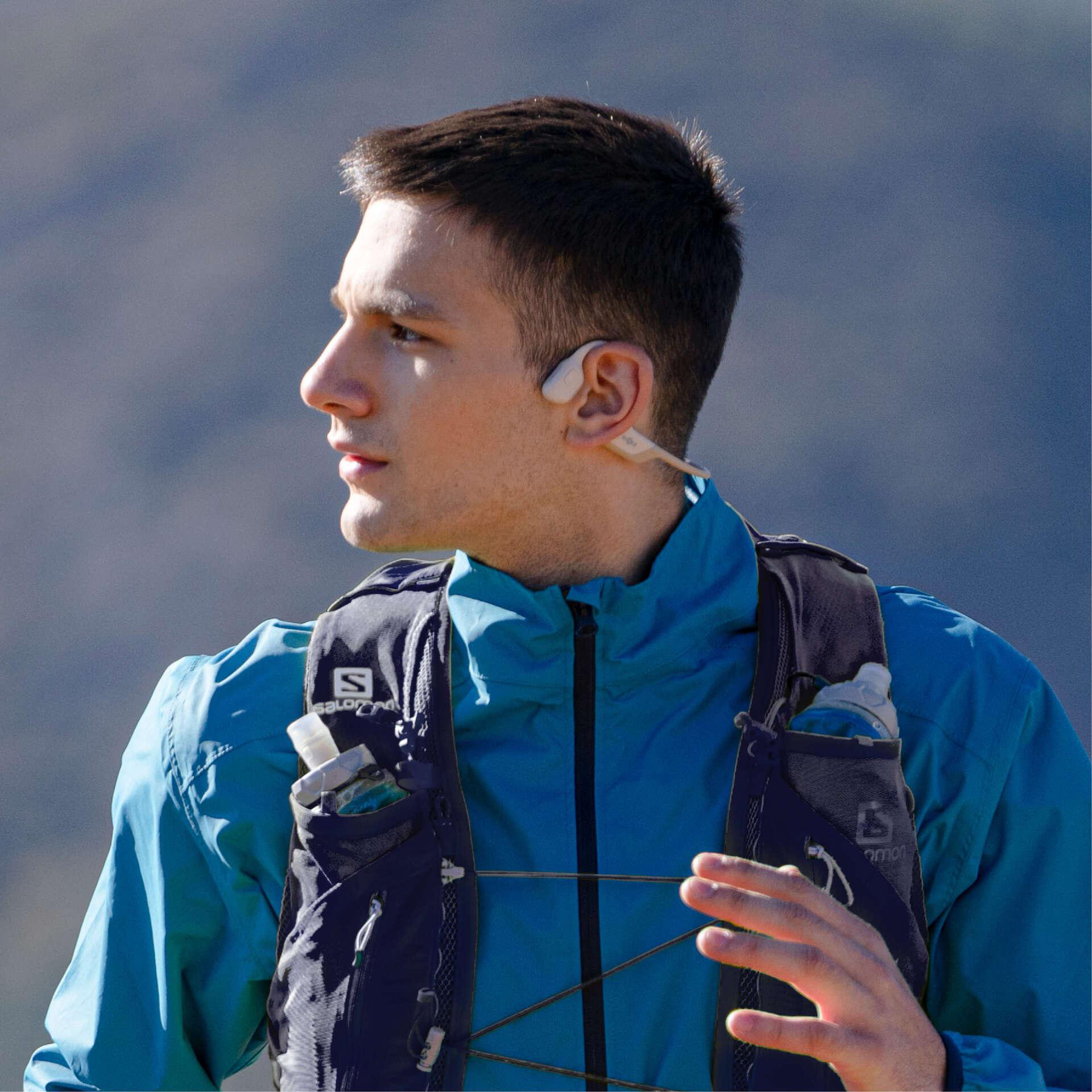 Ein Mann in einer blauen Jacke und Laufweste, der wegschaut. Er hat einen Ohrhörer und ist im Freien.