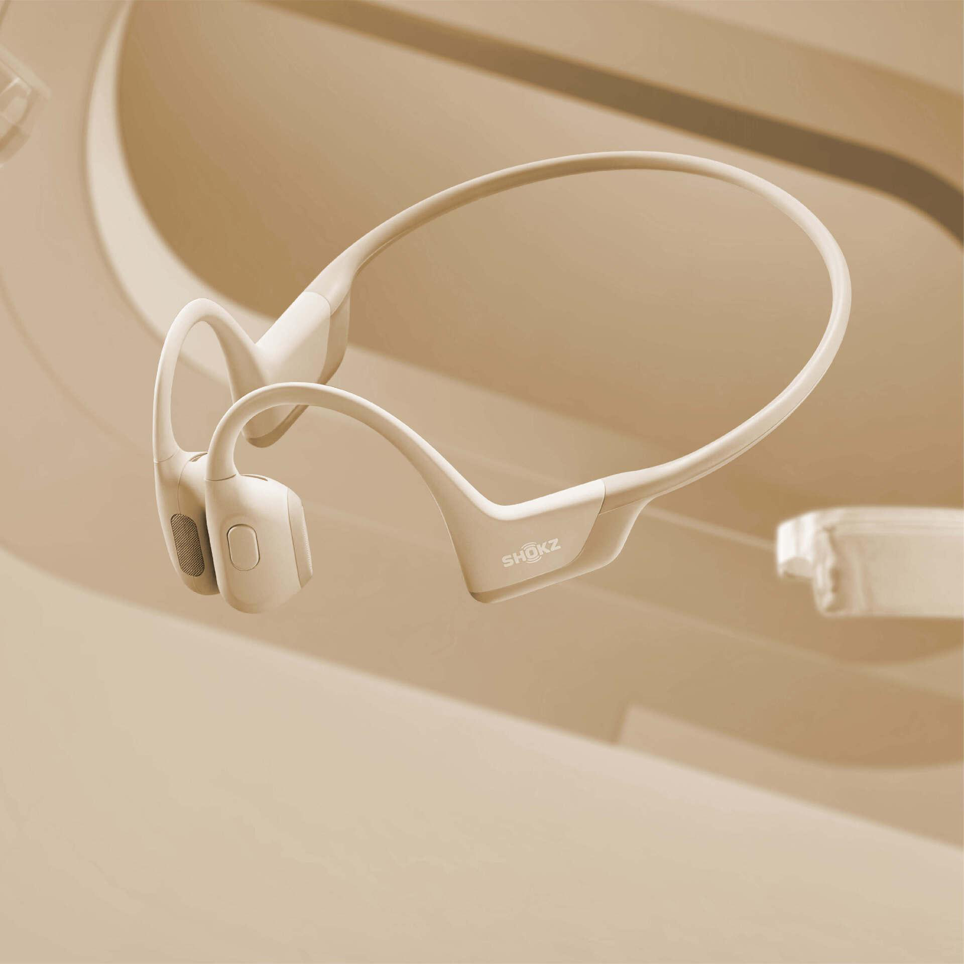 Knochenschallkopfhörer in Beige. Die Marke SHOKZ ist sichtbar. Der Hintergrund ist ein beiges, abstraktes Design.