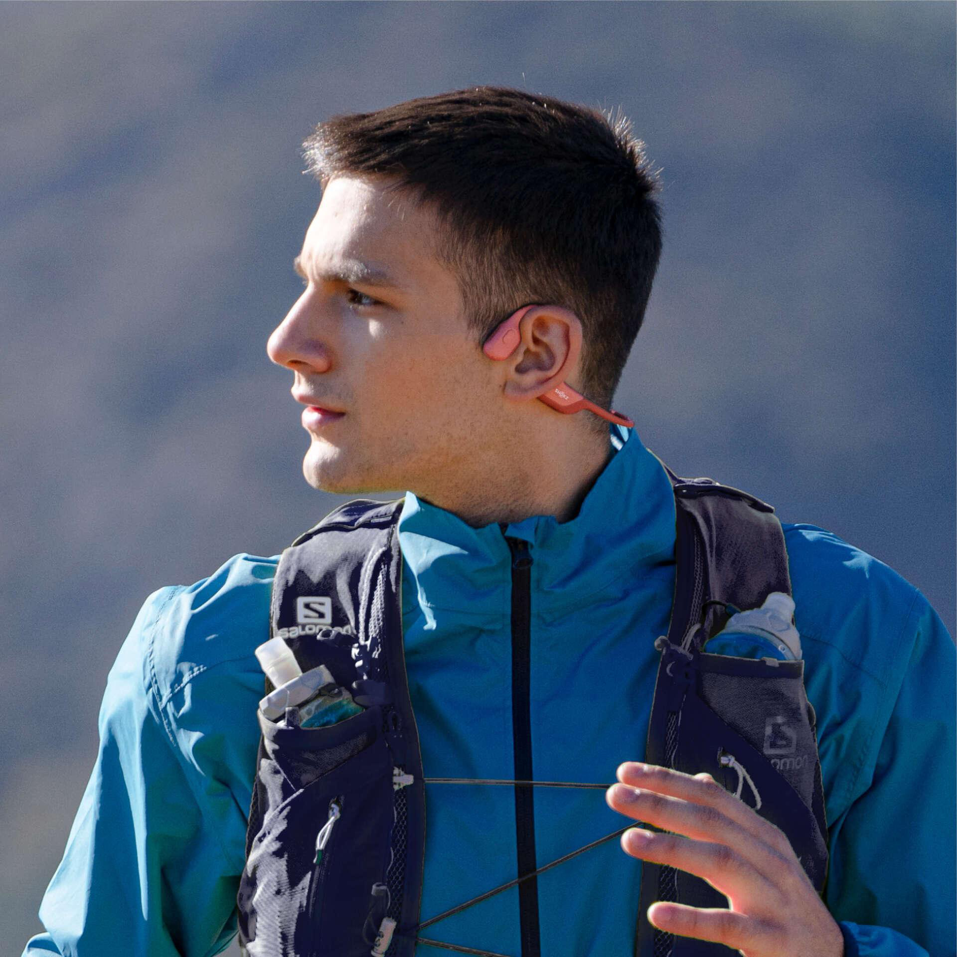 Mann mit rosa Kopfhörern, blauer Jacke, Laufweste. Berg Hintergrund. Blick nach links, teilweise sichtbare Hand rechts.