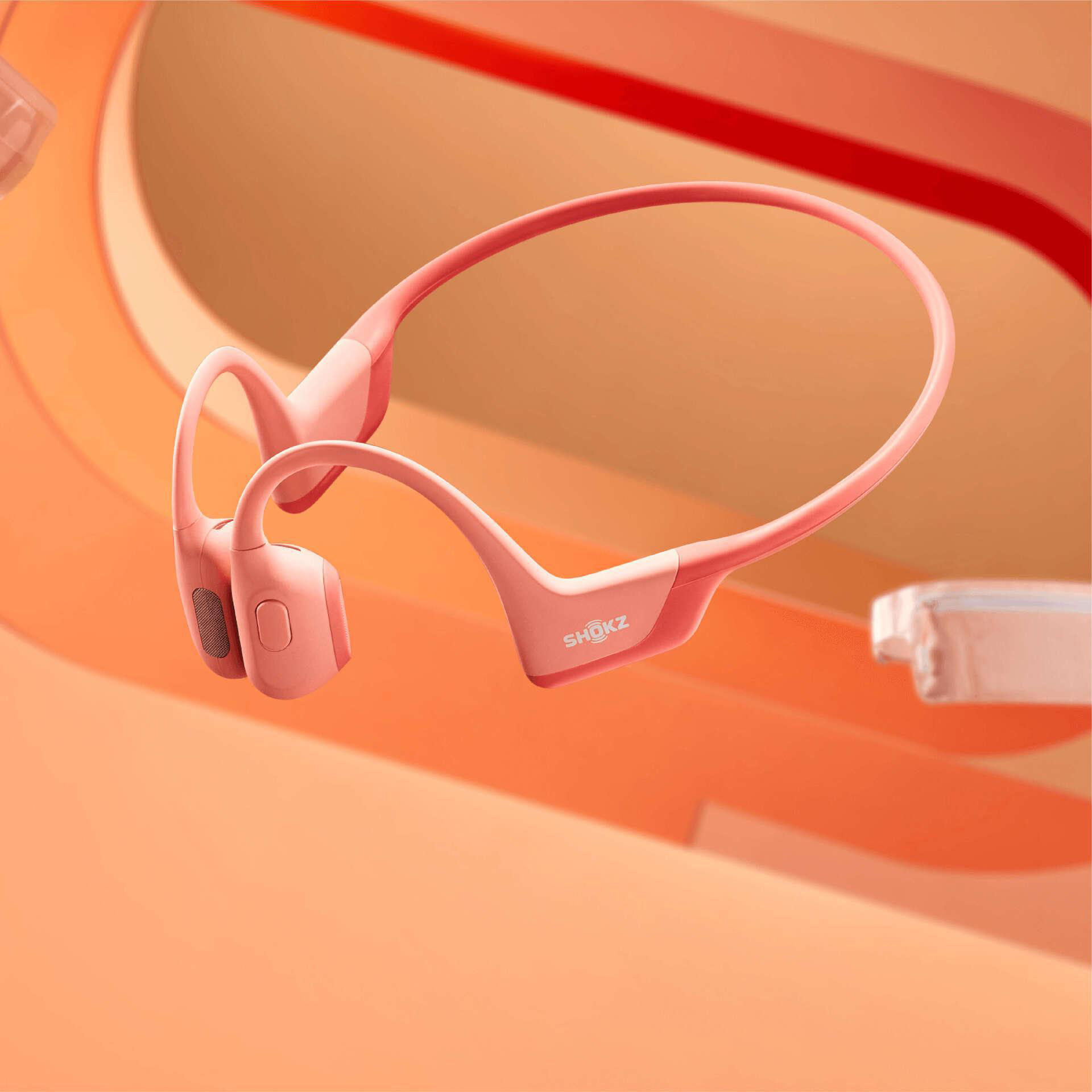 Rosa Shokz-Knochenschallkopfhörer auf orangefarbenem Hintergrund. Die Kopfhörer sind im Fokus, der Markenname ist sichtbar.