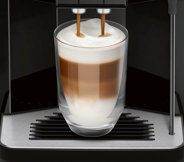 Kahve makinesi, bir bardağa kahve döküyor. Bardak şeffaf ve katmanlı bir latte görünümüne sahip.