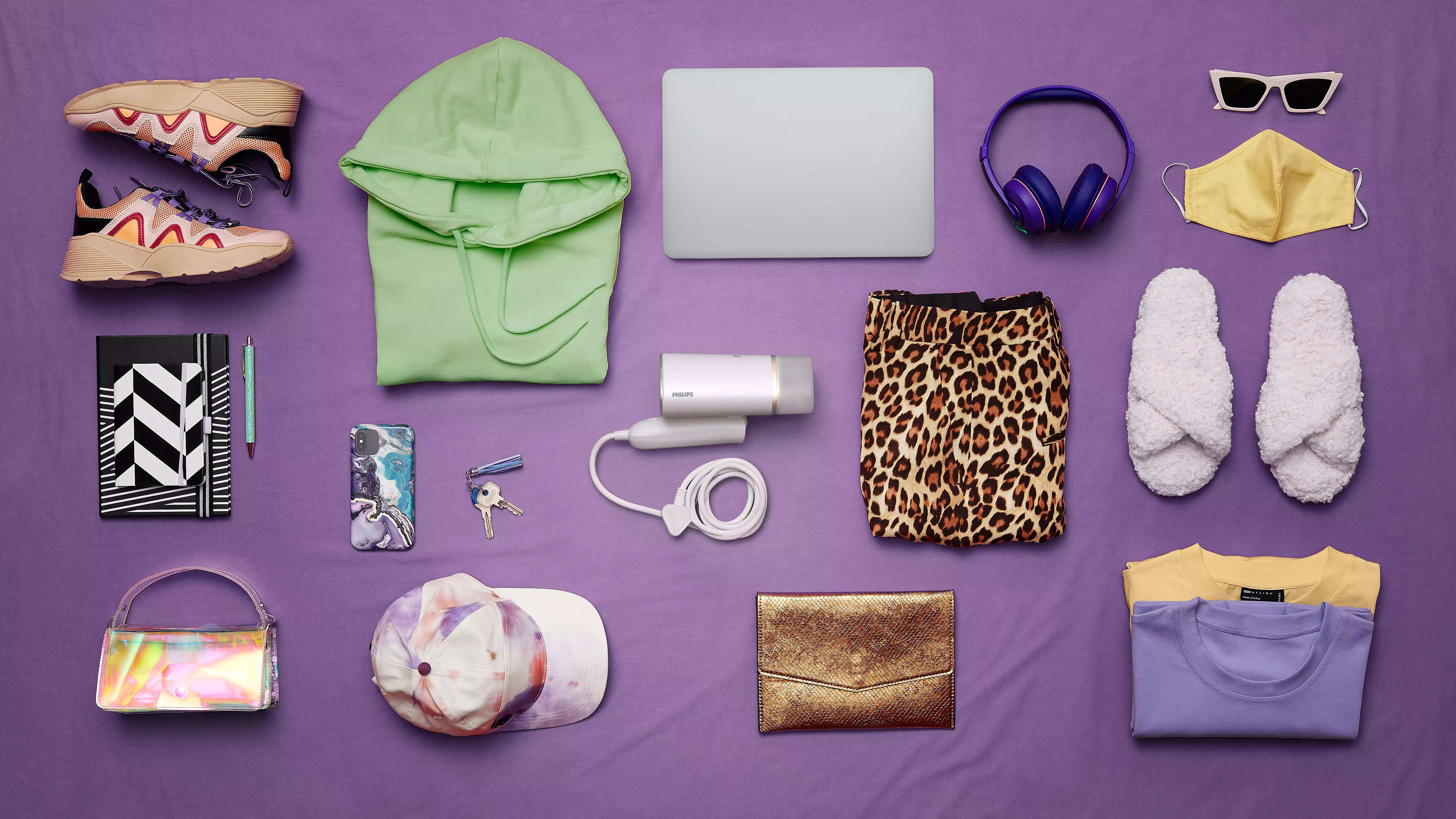 Divers objets disposés sur une surface violette : chaussures, sweat à capuche, ordinateur portable, accessoires.