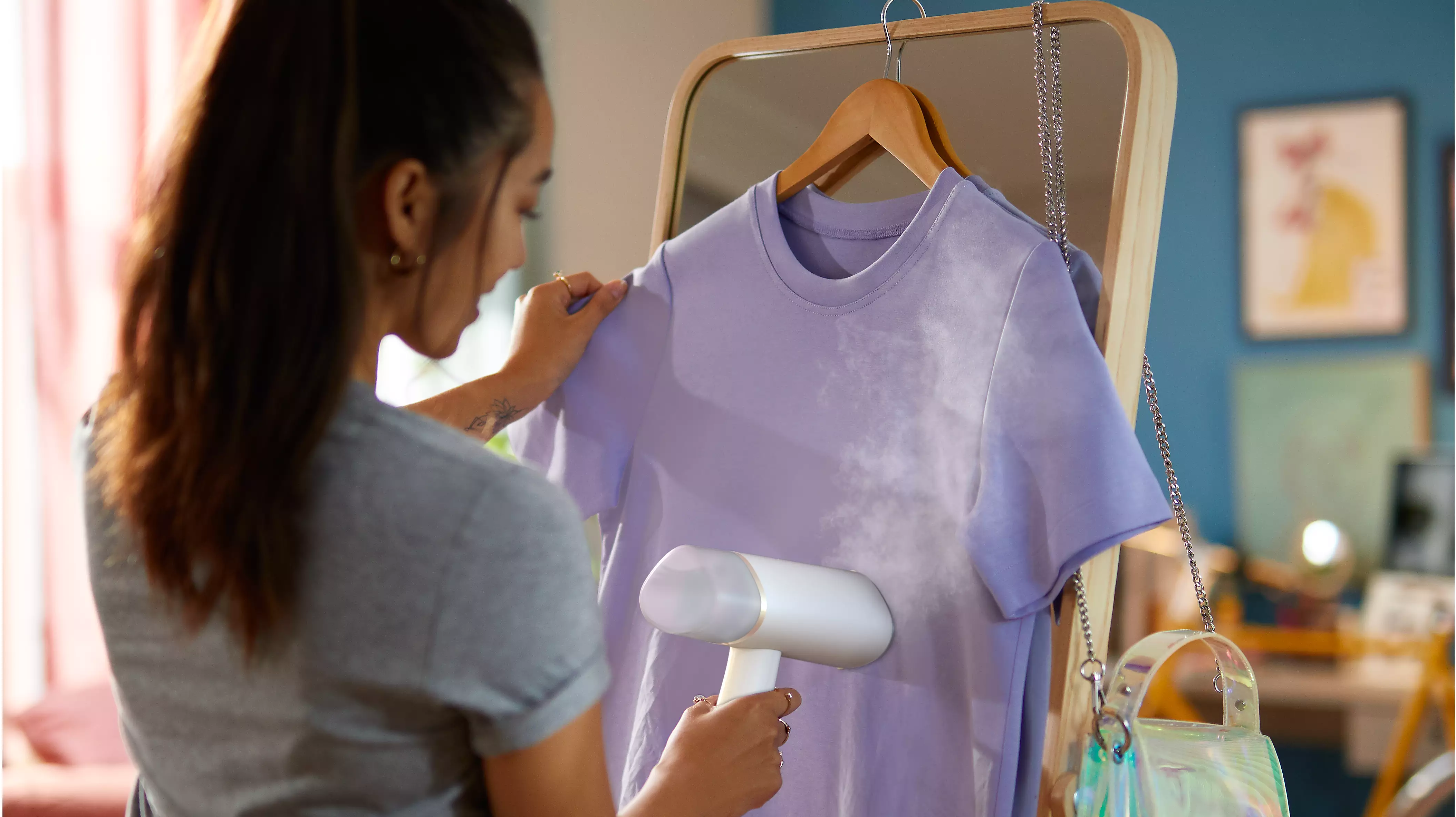 Une femme repassant un t-shirt violet avec un défroisseur à main, près d'un miroir et d'un sac à main.