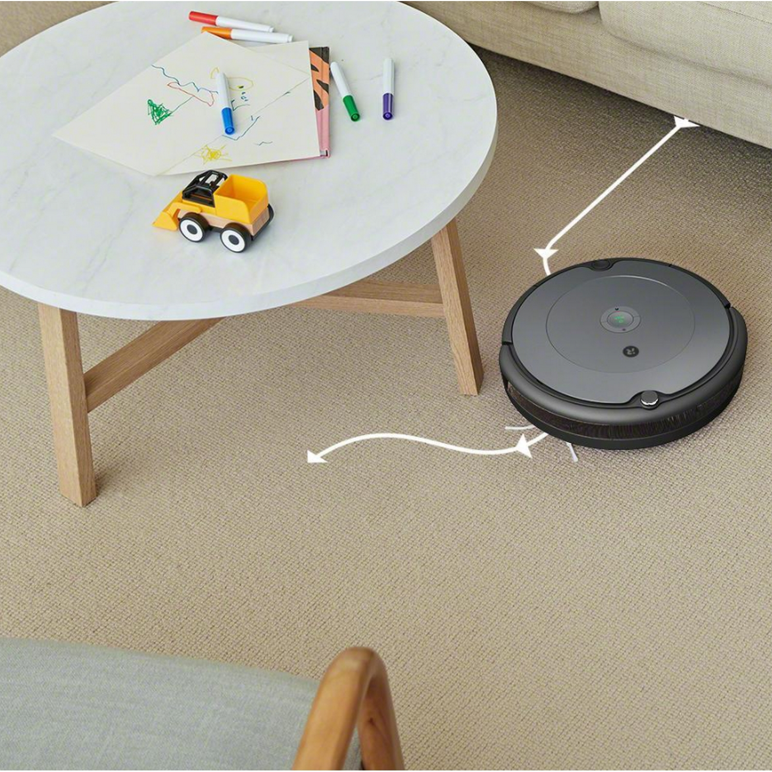 Una mesa blanca con dibujo y juguete en primer plano. Un robot aspirador en el suelo.