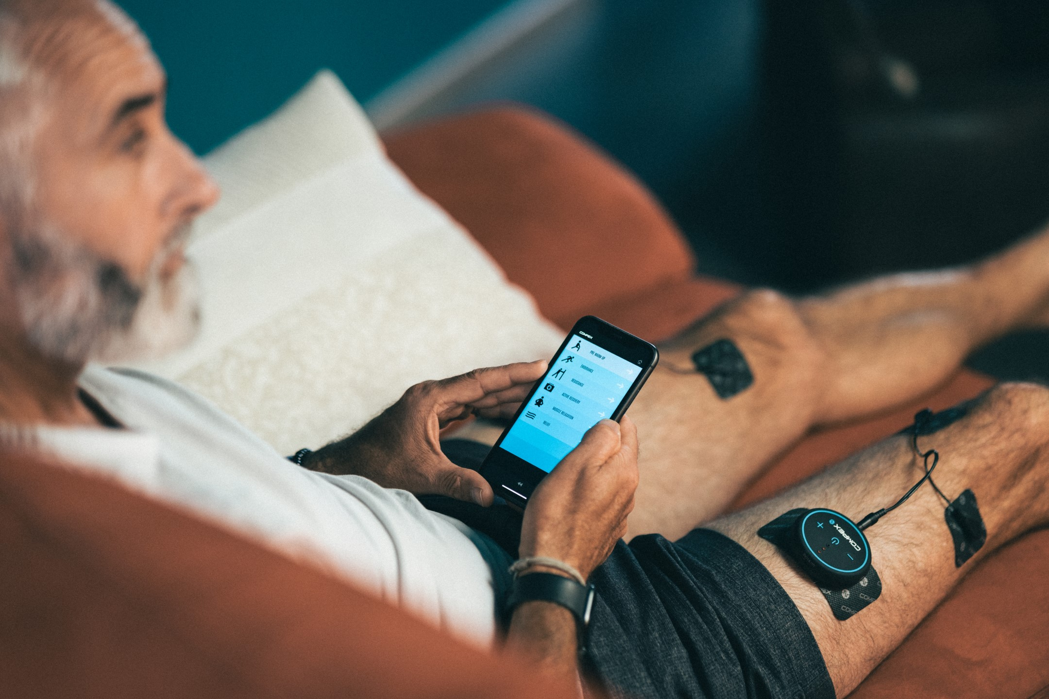 Mann auf Couch, Smartphone nutzend, Elektroden an Beinen.