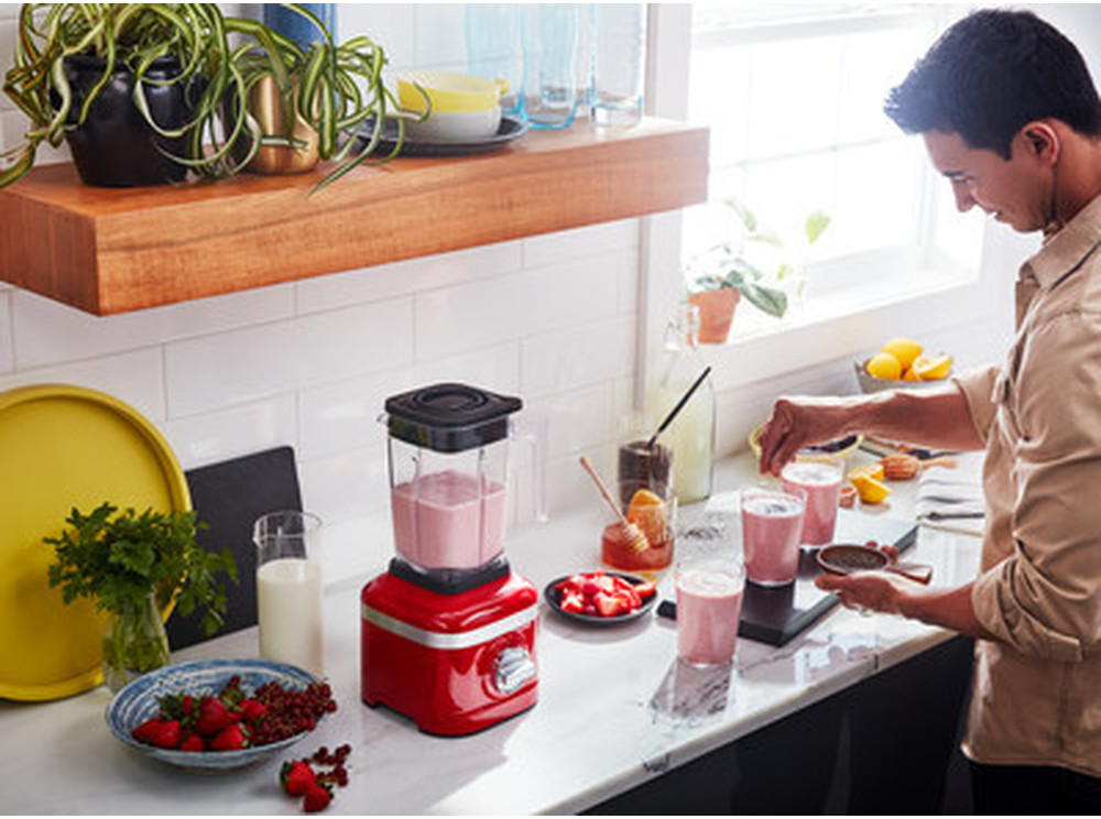 Un homme prépare des smoothies dans une cuisine, mixeur, fraises et verres sur le comptoir.