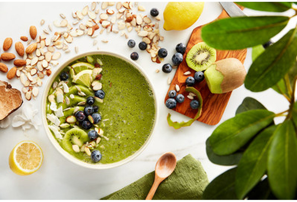 Bovenaanzicht van een groene smoothiekom met fruit, noten en een houten lepel op wit.