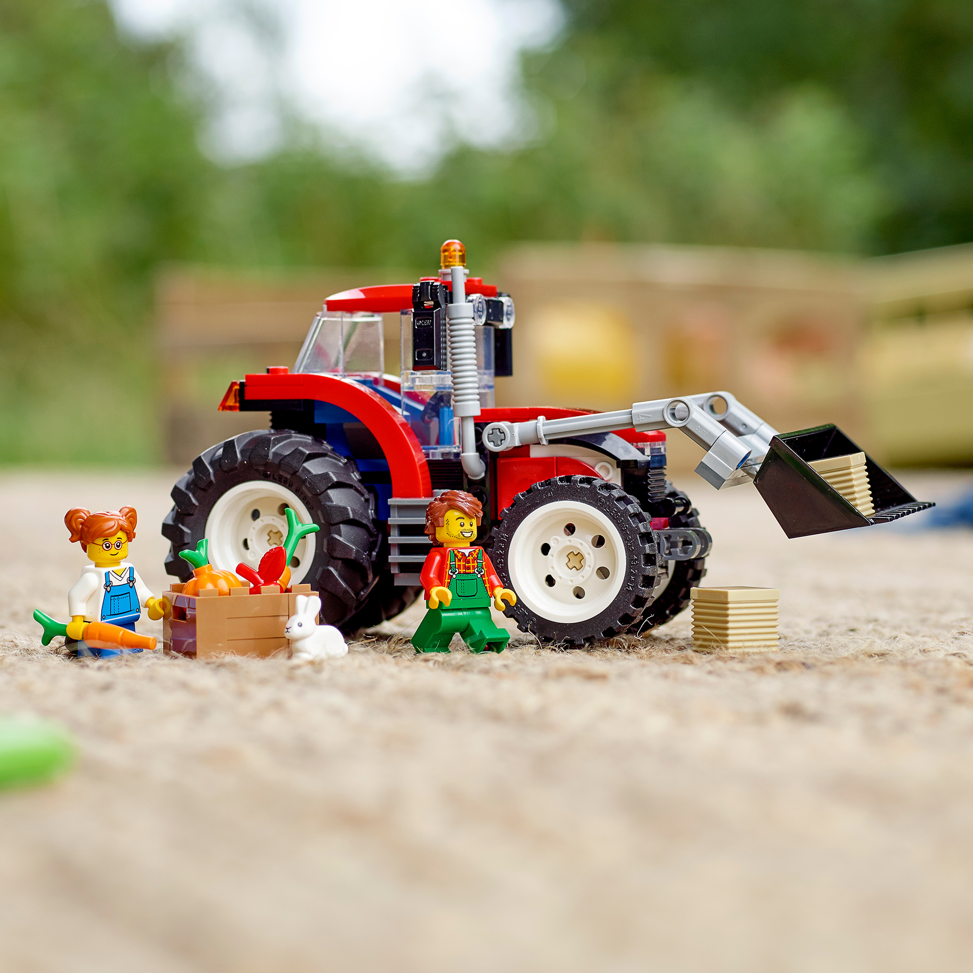 Ein roter Lego-Traktor mit Frontlader, zwei Minifiguren, einer Gemüsekiste und einem weißen Hasen auf Sand.