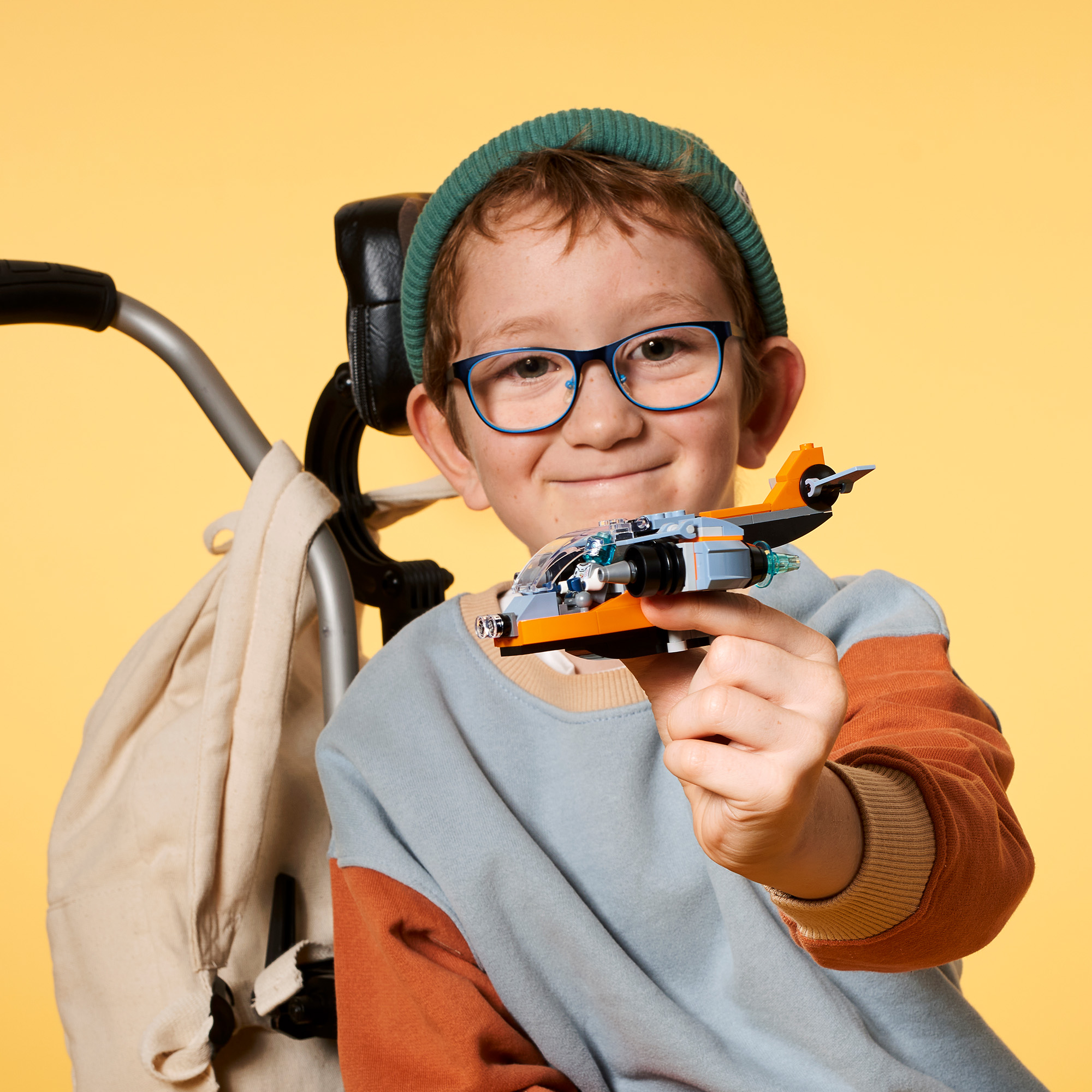 Junge hält ein Spielzeugflugzeug, trägt Brille und Mütze, gelber Hintergrund.