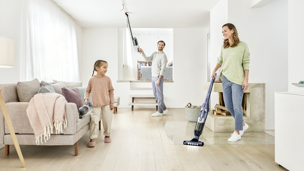 Family cleaning the house. A woman is vacuuming, a man uses a stick vacuum, and a girl holds one.