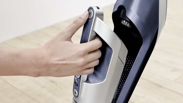 A hand is shown pressing a button on a dark blue and silver vacuum cleaner.