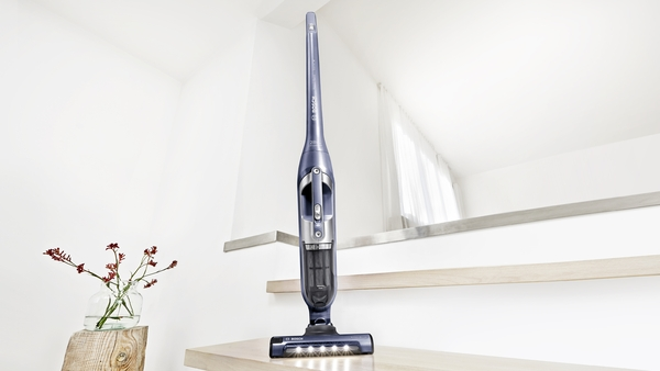 A cordless vacuum cleaner is standing on a staircase. Red flowers in a vase are in the background.