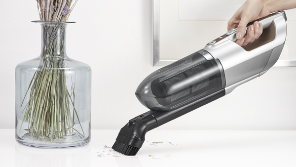 A hand holding a small vacuum cleaner over a surface with dried flowers in a vase.