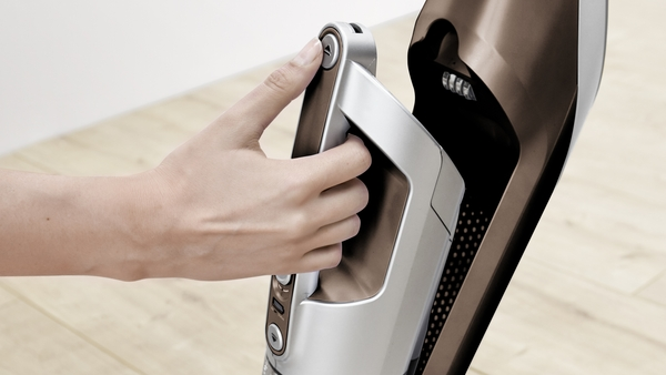 A hand operates a brown and silver vacuum cleaner. The floor is light wood.