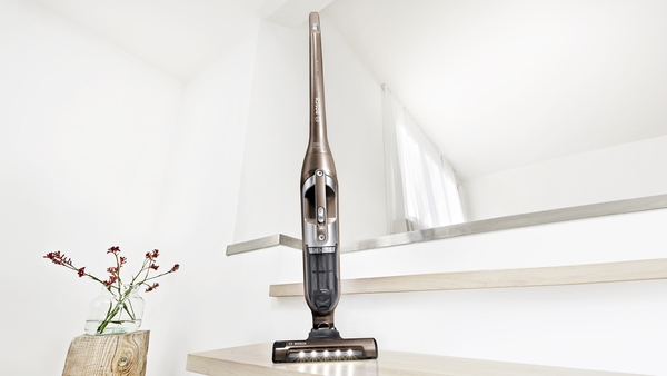 A cordless vacuum cleaner on a wooden step with a small flower vase in the background.