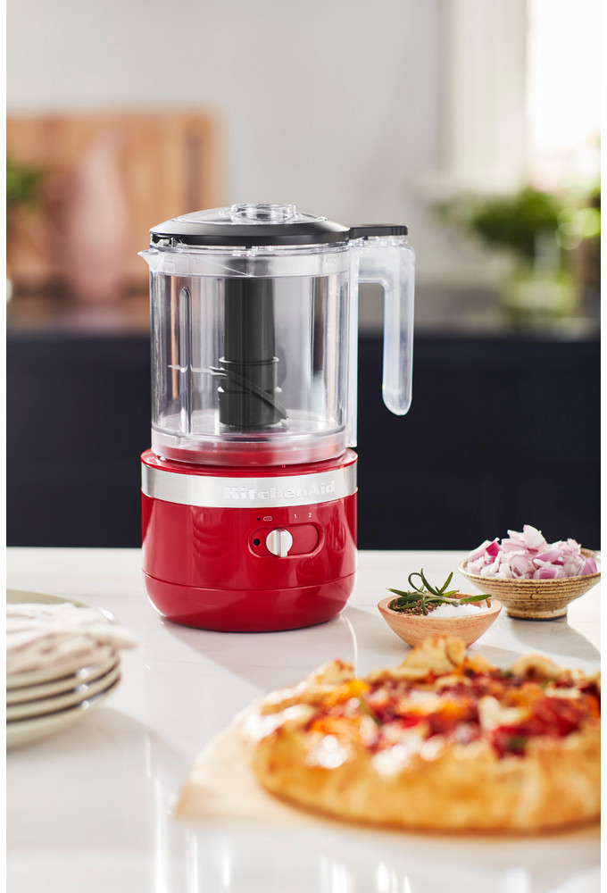 Rode KitchenAid foodchopper op een aanrecht met rode basis. Een taart en kommen met eten staan op de voorgrond.