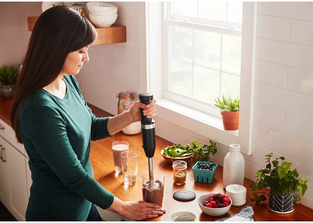 Woman blends smoothie with immersion blender. Kitchen setting with ingredients.