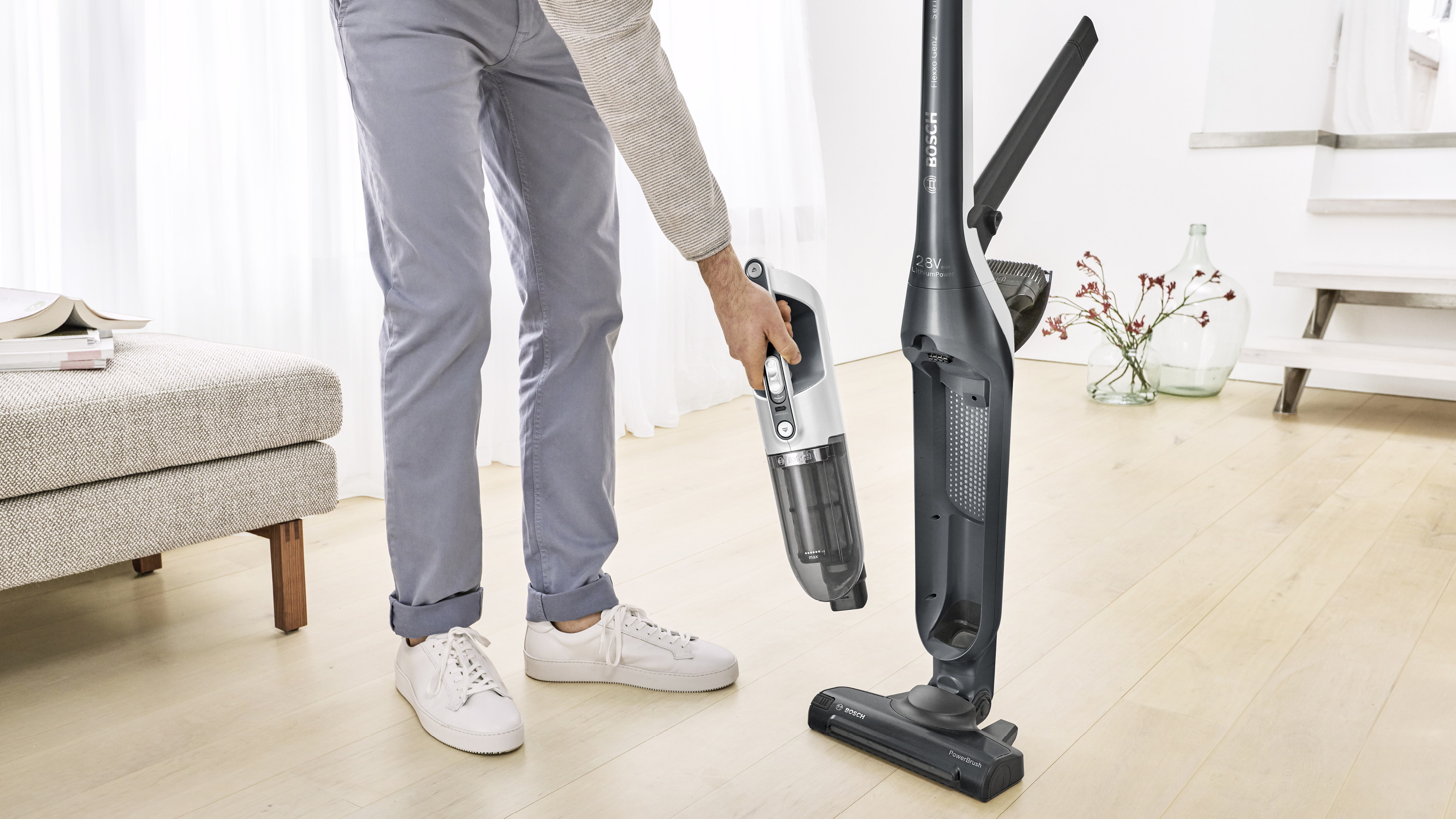 Mann hält einen weißen und grauen kabellosen Staubsauger. Rechts steht ein Staubsauger.