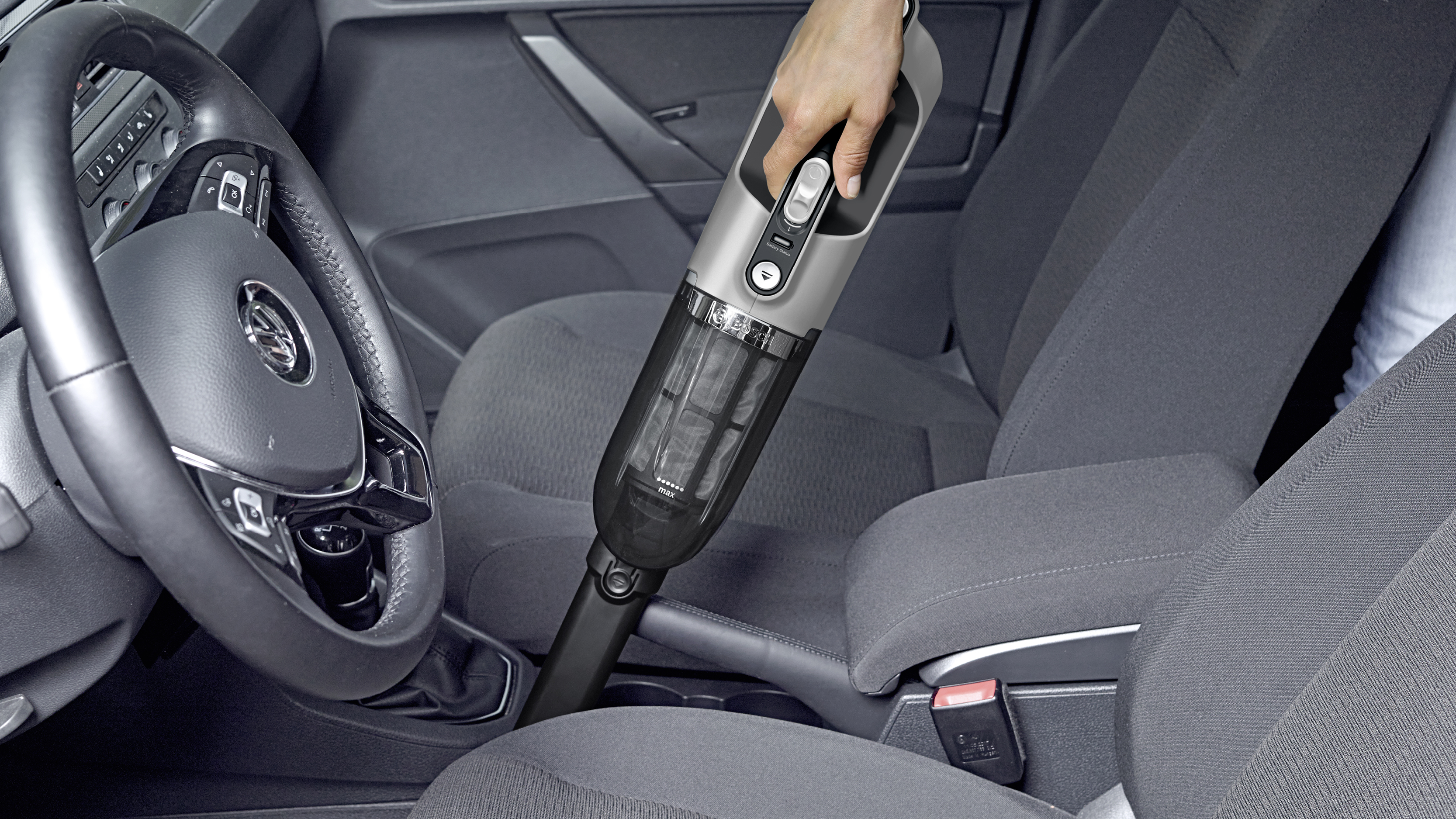 Person reinigt Autoinnenraum mit einem Handstaubsauger. Autositz und Lenkrad sind sichtbar.