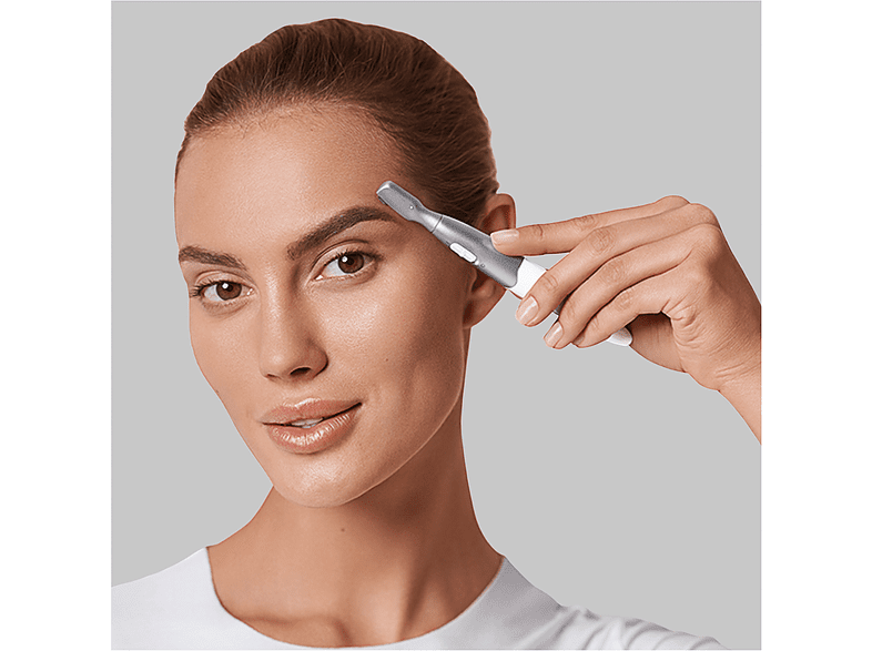 A woman using an eyebrow trimmer. She holds the trimmer to her eyebrow, looking at the camera.