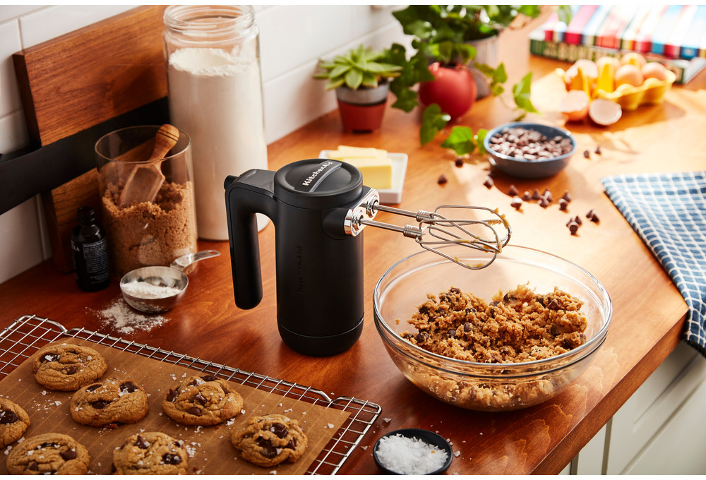 A black KitchenAid mixer and bowl with ingredients, cookies, and baking supplies on a wooden counter.