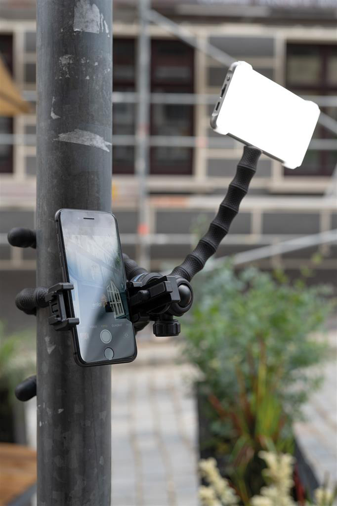 Egy telefon és egy lámpa egy oszlopra van szerelve. A telefon egy épületet mutat. A háttérben építkezés és növények láthatók.