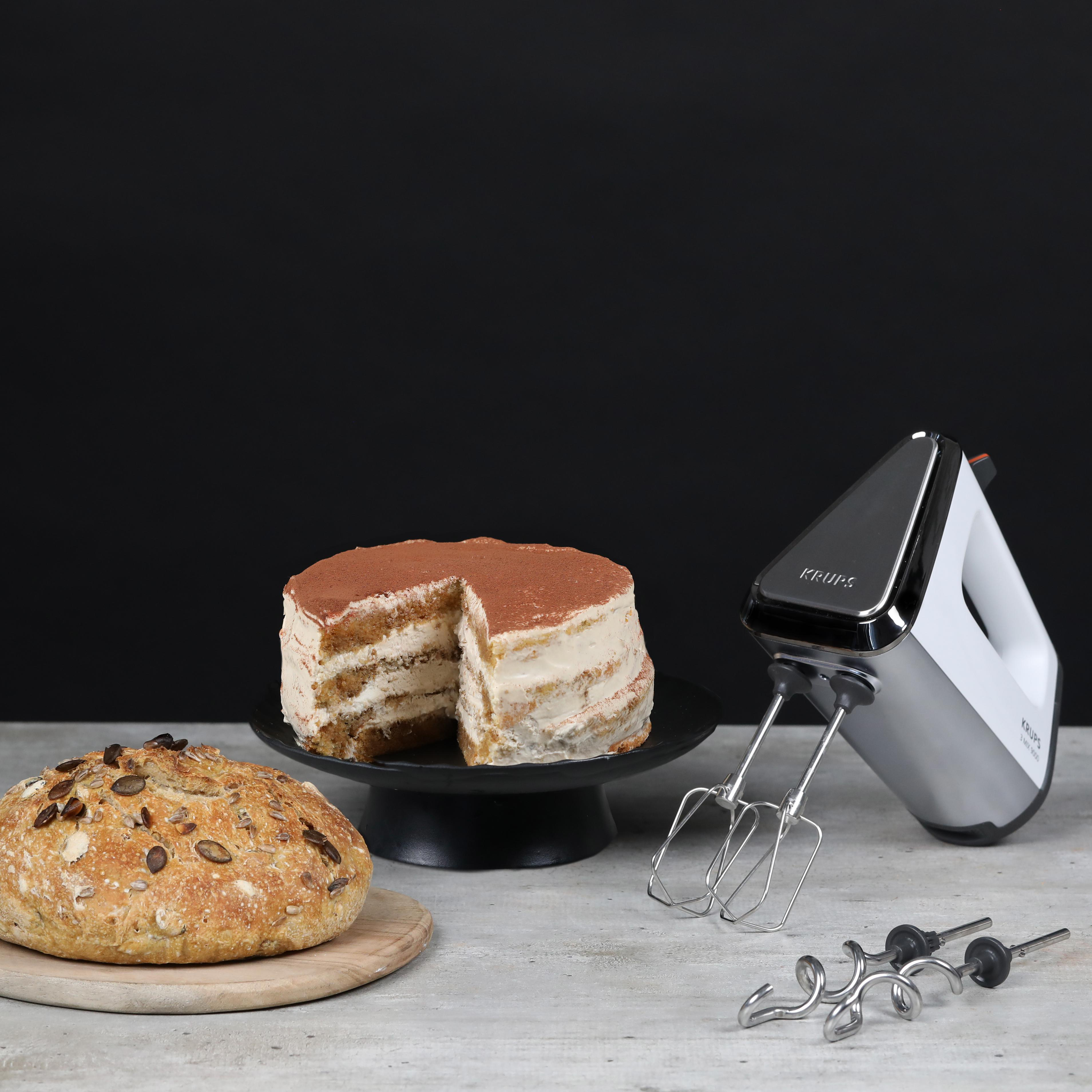 Ein Tiramisu-Kuchen auf einem Ständer, ein Brotlaib und ein Handmixer. Alles auf hellfarbener Oberfläche.