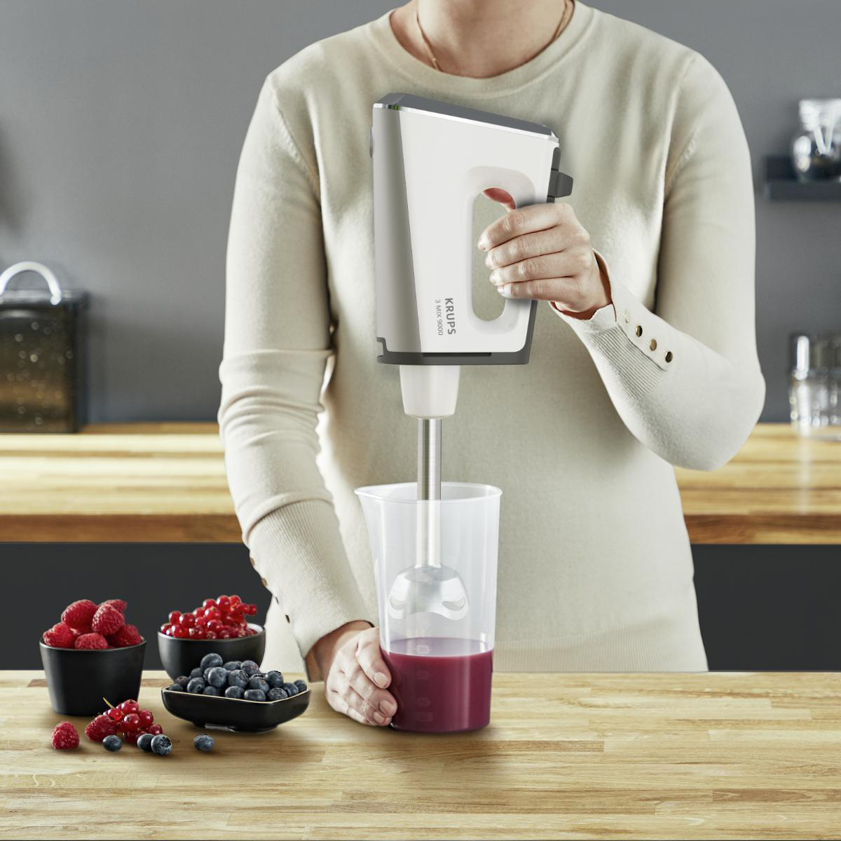 Person verrührt Zutaten mit einem Handmixer in einem transparenten Behälter. Beeren sind sichtbar.
