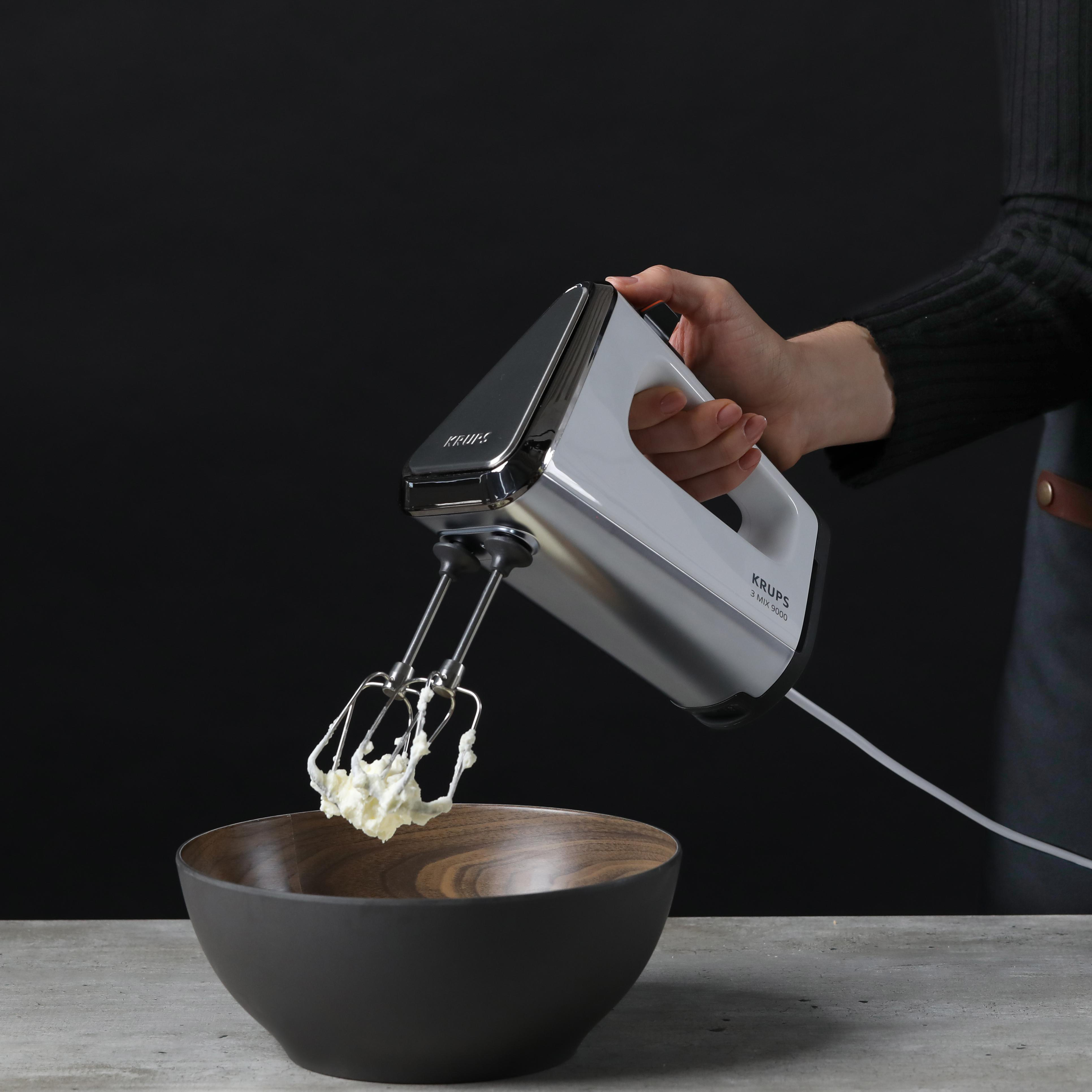 Eine Hand, die einen silbernen Handmixer über einer Holzschüssel mit Sahne hält.