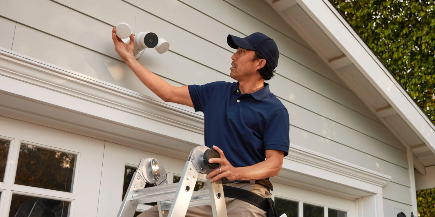Man on a ladder is installing a security light on a house exterior. It is daytime.