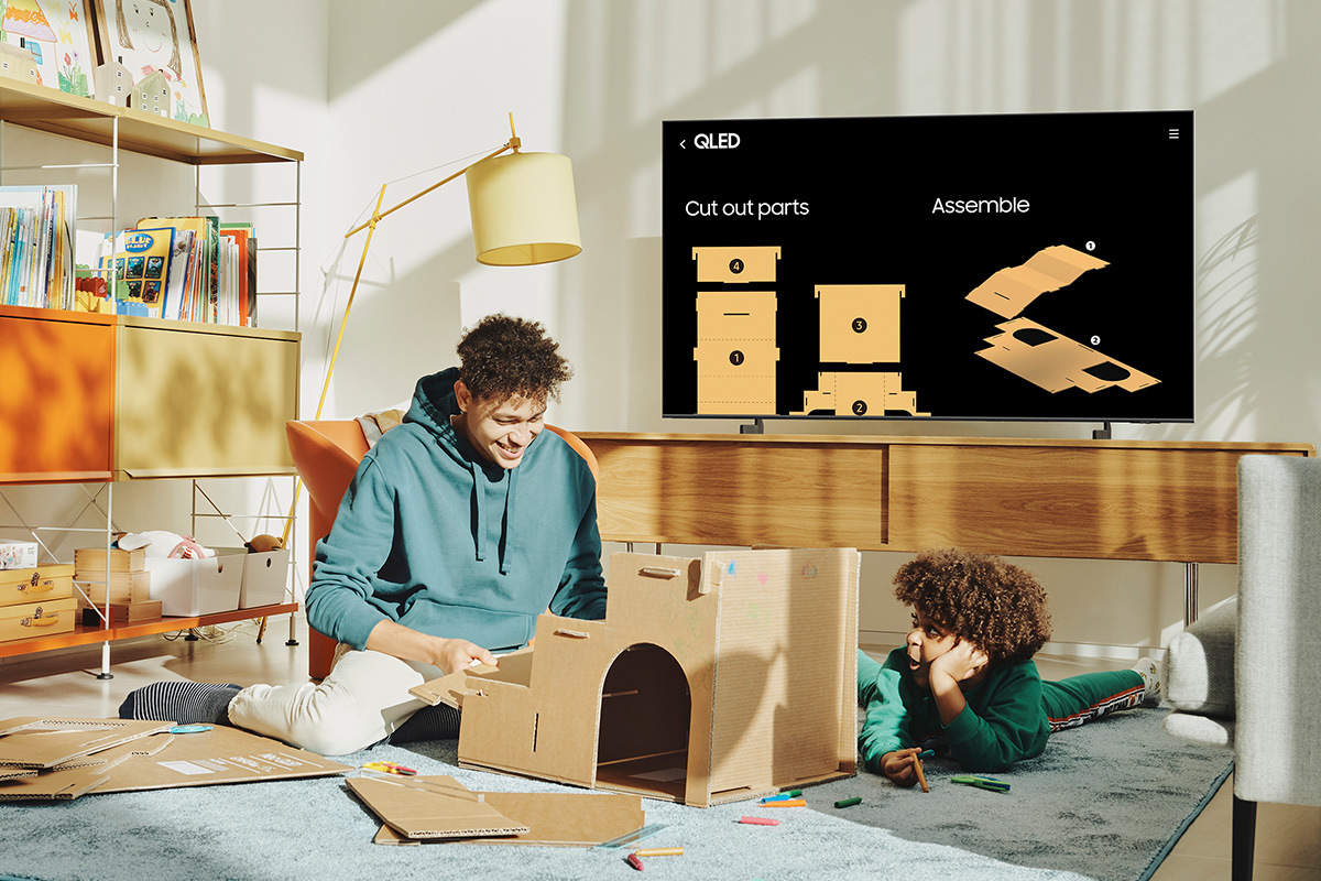 Deux enfants construisant un château en carton. Une télévision affiche des instructions de découpe. Salon avec une lampe jaune.