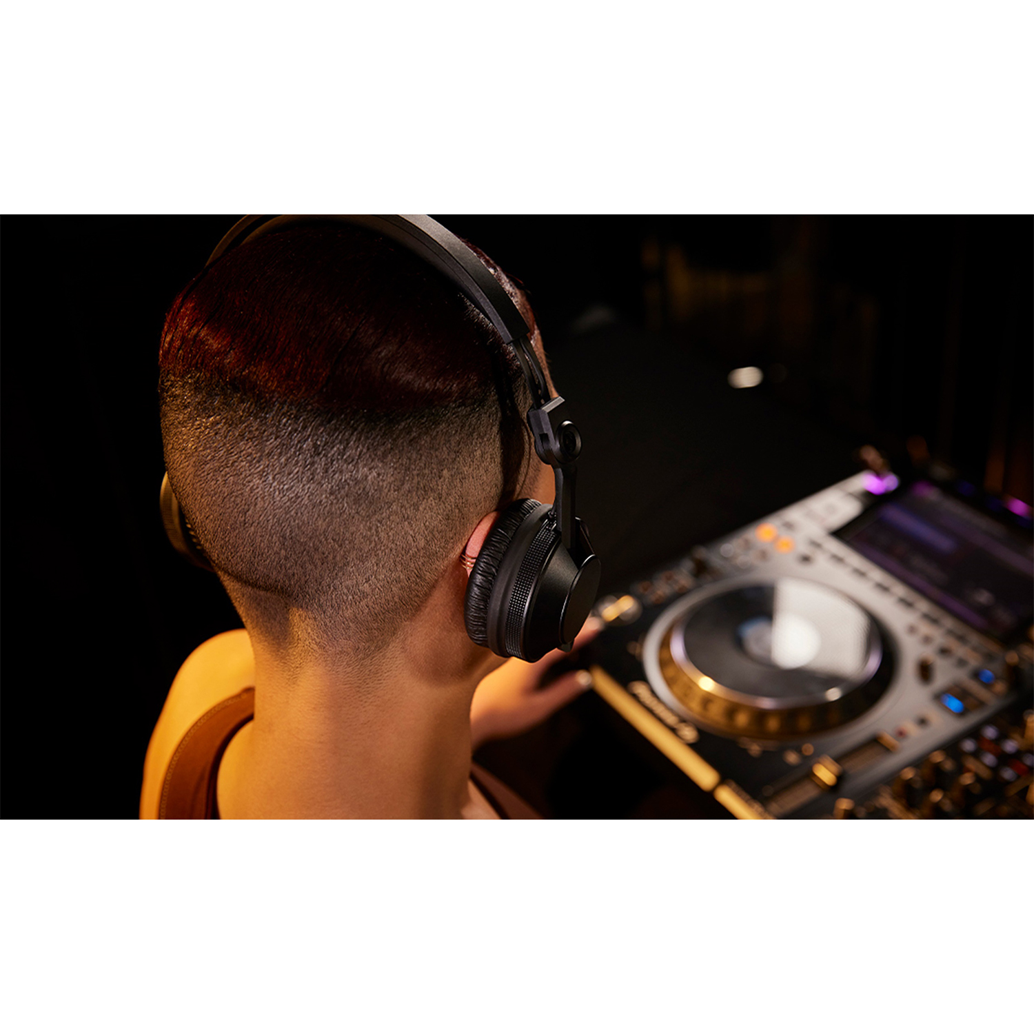 Una persona con taglio corto indossa cuffie da DJ nere, con attrezzatura da DJ dietro.