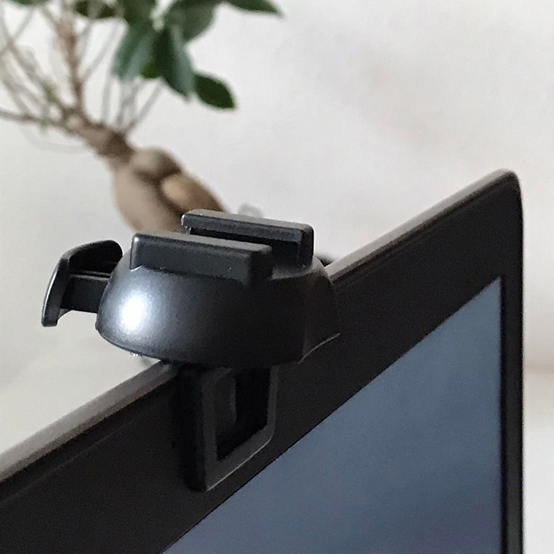 Egy fekete webkamera-fedél van egy laptop képernyőjére rögzítve. Egy elmosódott növény a háttérben, fehér fal előtt.