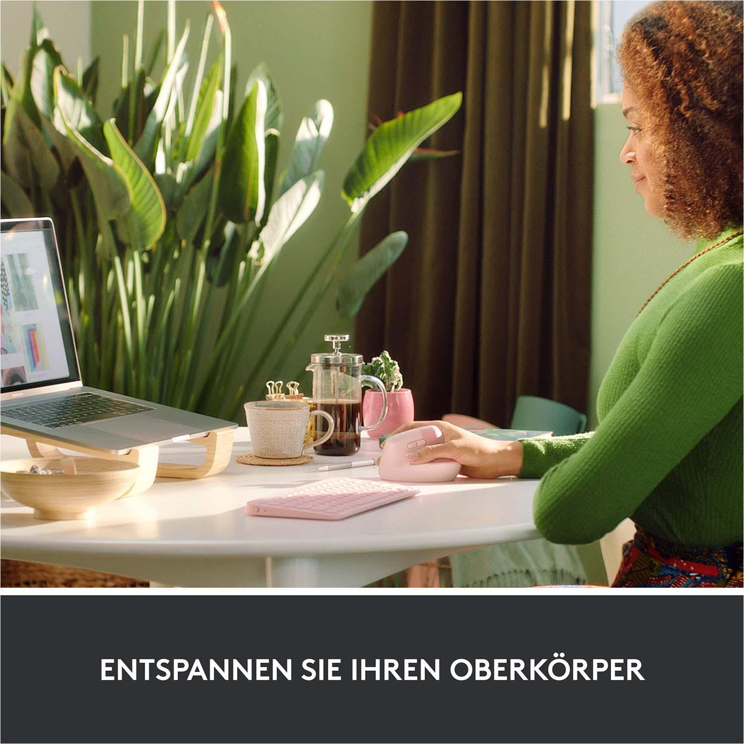 Frau arbeitet am Laptop, rosa Maus und Tastatur auf Tisch. Pflanzen im Hintergrund.