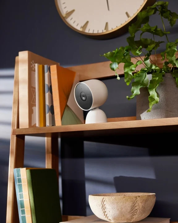 Een witte beveiligingscamera op een houten plank met boeken en een plant.