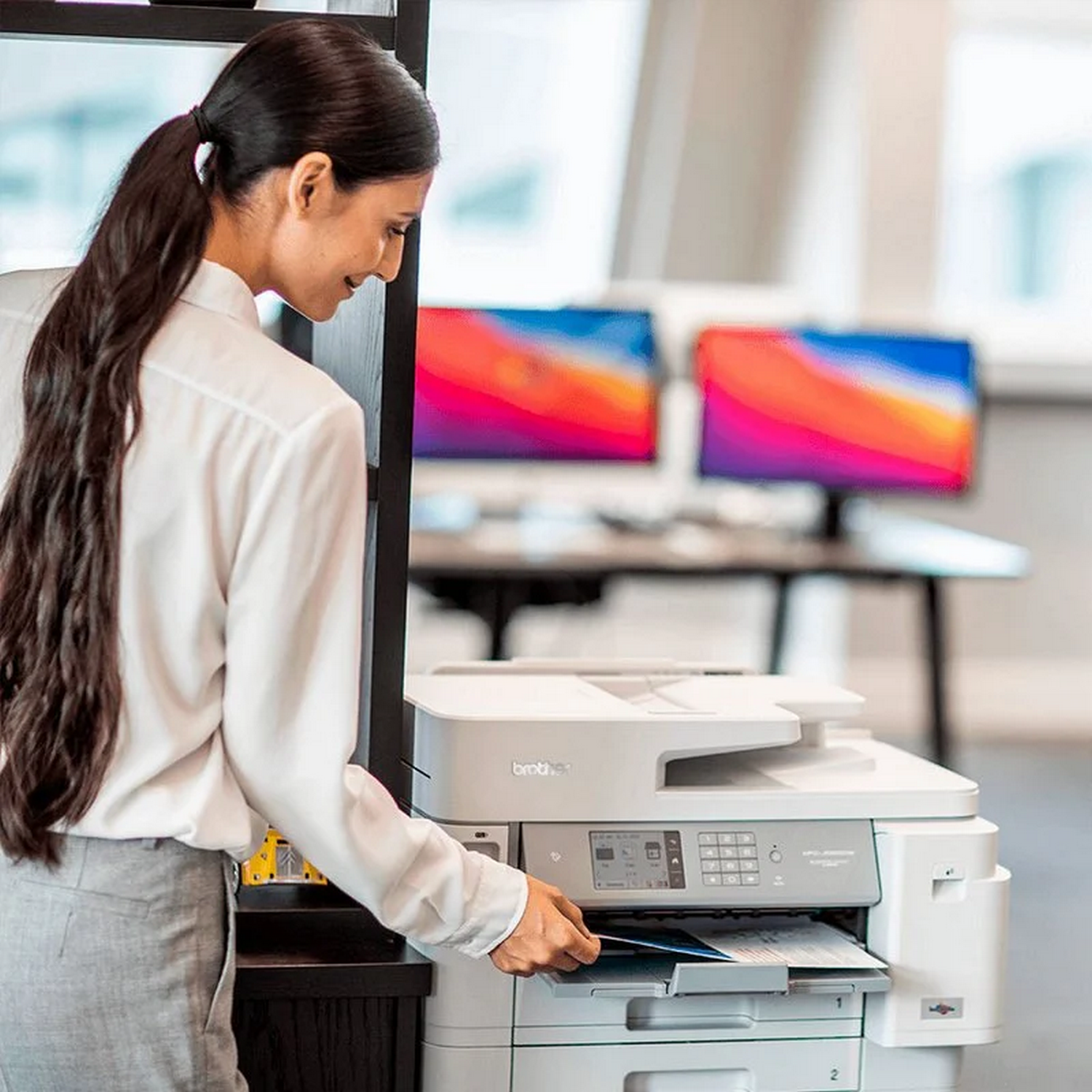 Una mujer con camisa blanca usa una impresora Brother, en un entorno de oficina. Dos monitores en el fondo.