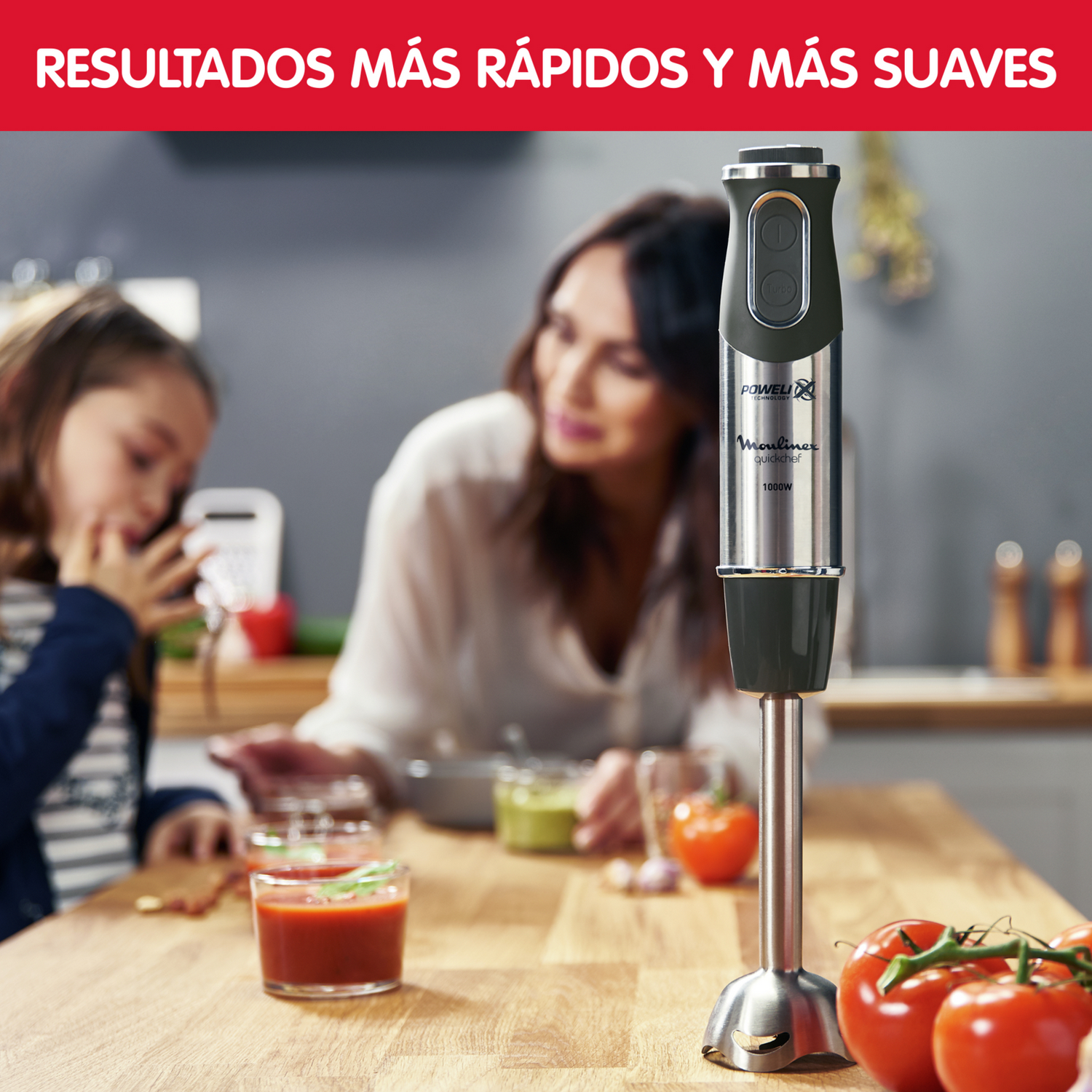 Una batidora de mano Moulinex siendo utilizada por alguien mientras otro observa. Los tomates están en primer plano.