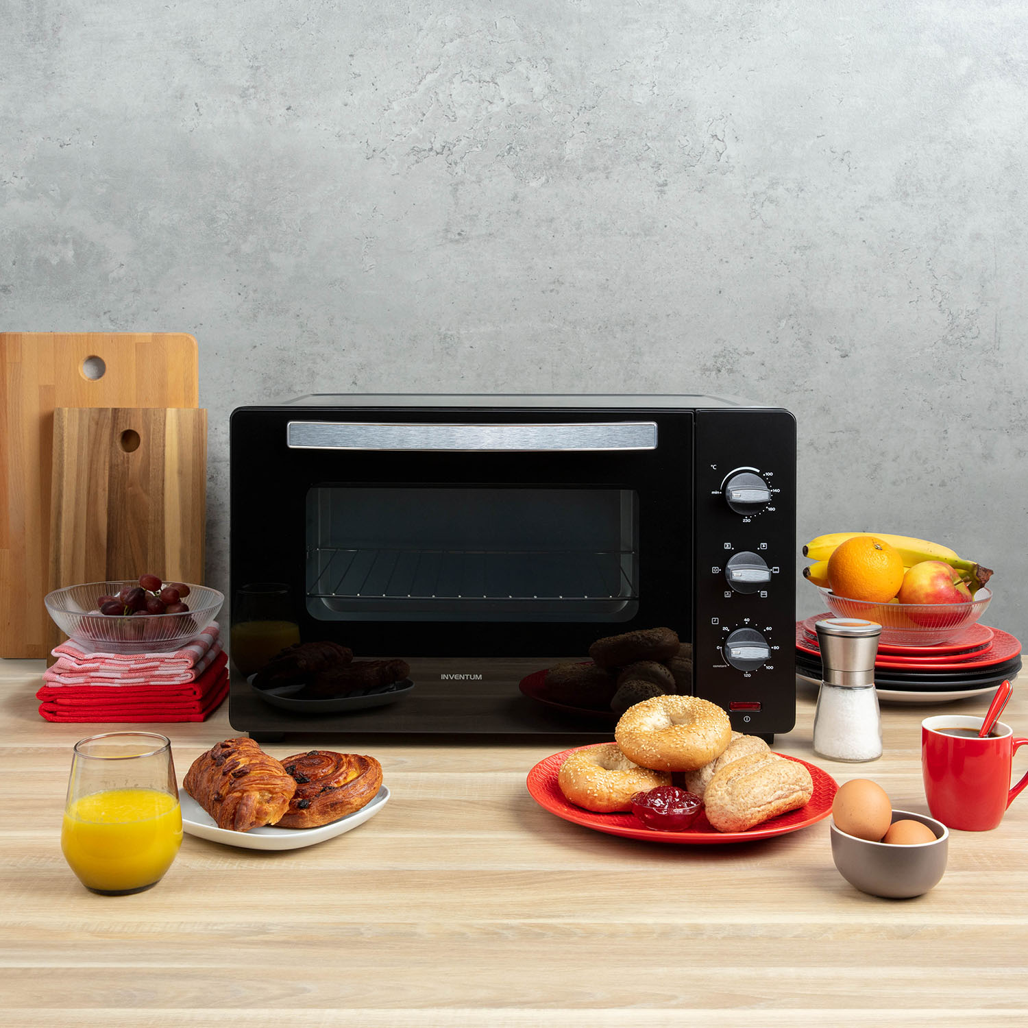 A black oven with food items on a table: croissants, bagels, fruit, eggs, and juice, on a wood surface.