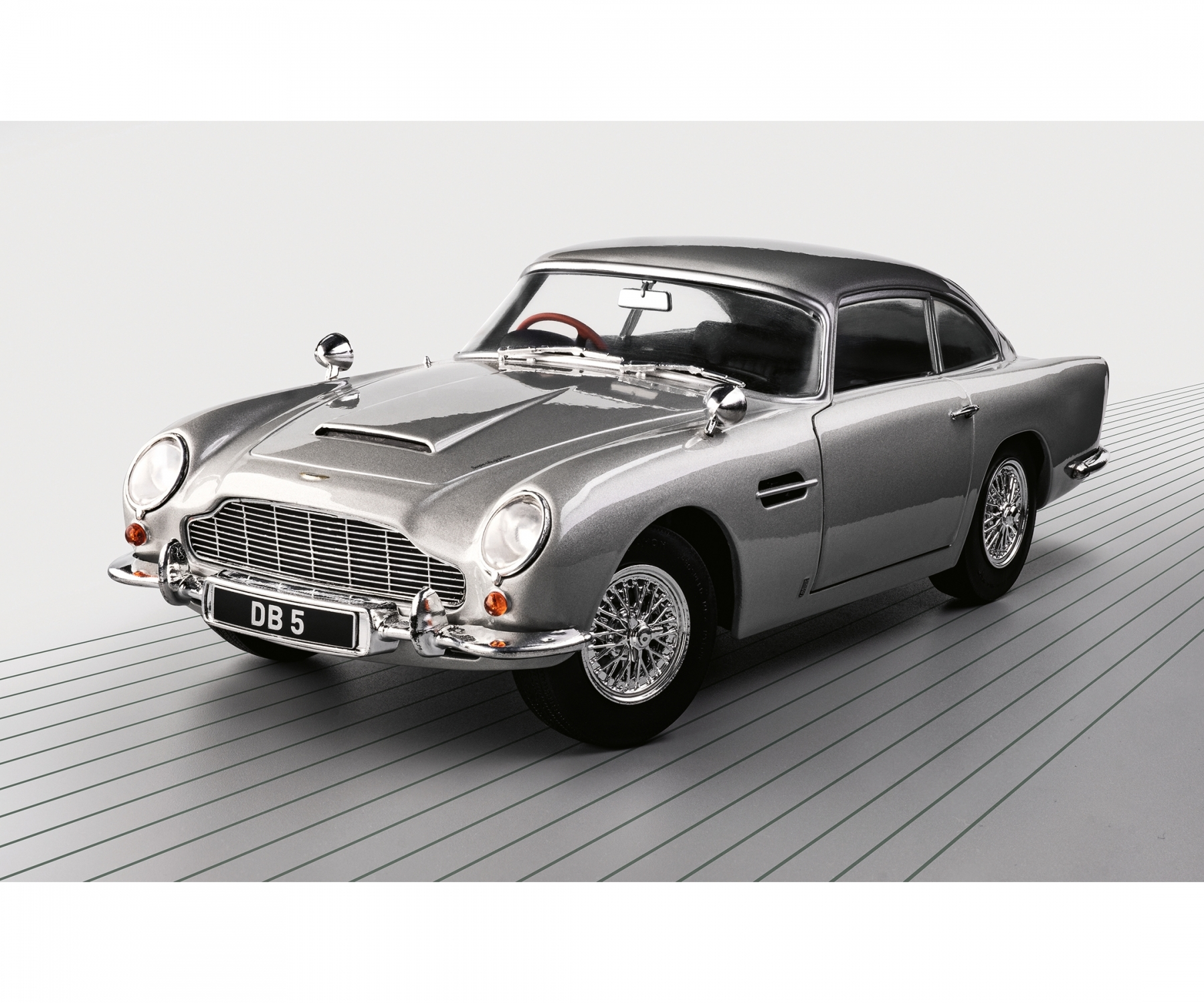 Ein silberner Oldtimer, ein DB5, steht auf einer grauen Oberfläche mit Linien. Das Auto hat Chromdetails und ein schwarzes Nummernschild.